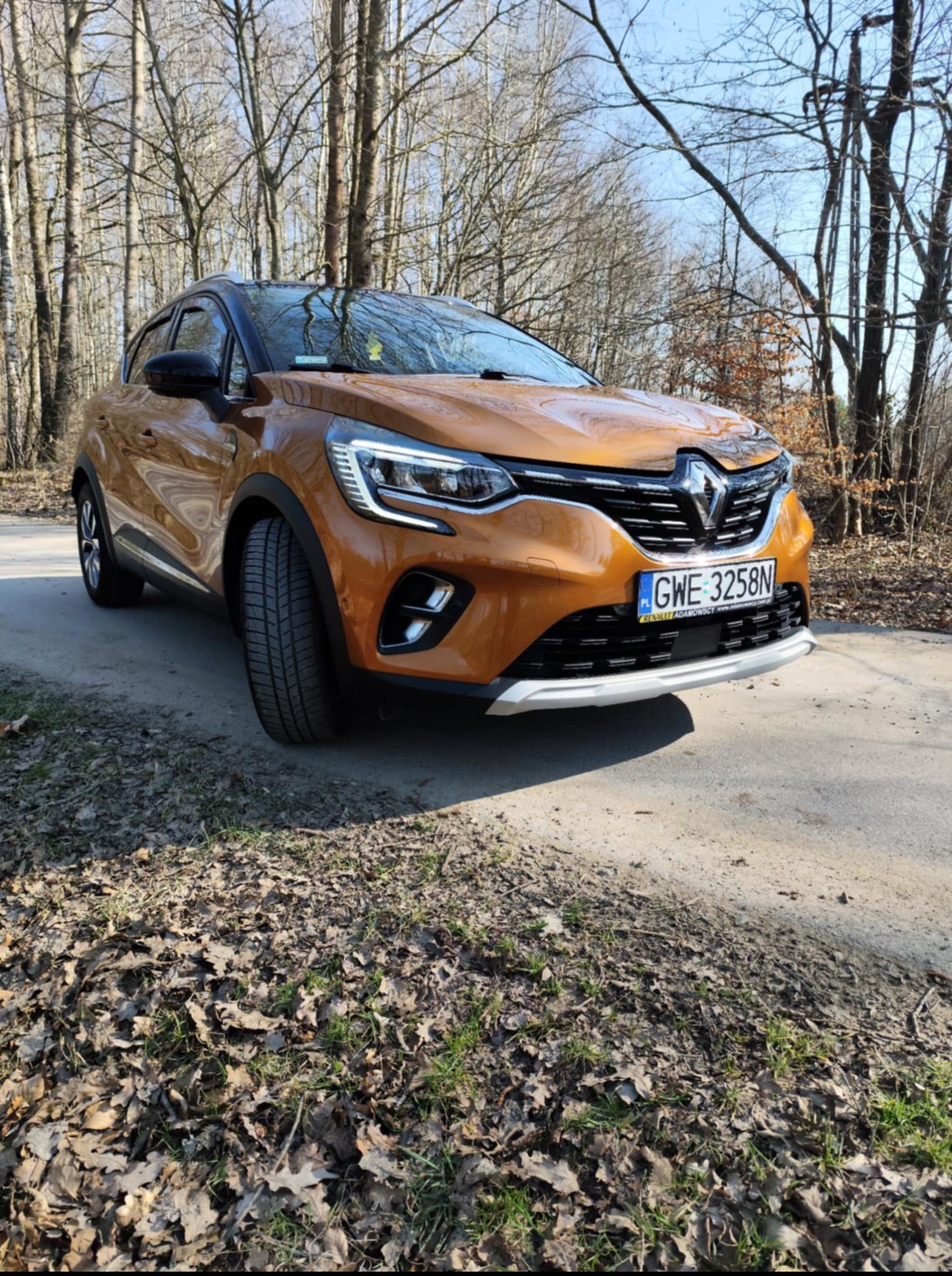 Pomarańczowy Renault Captur na leśnej drodze, widok z przodu, tablica rejestracyjna GWE 3258N, jesienne liście na poboczu, drzewa w tle, słoneczny dzień.