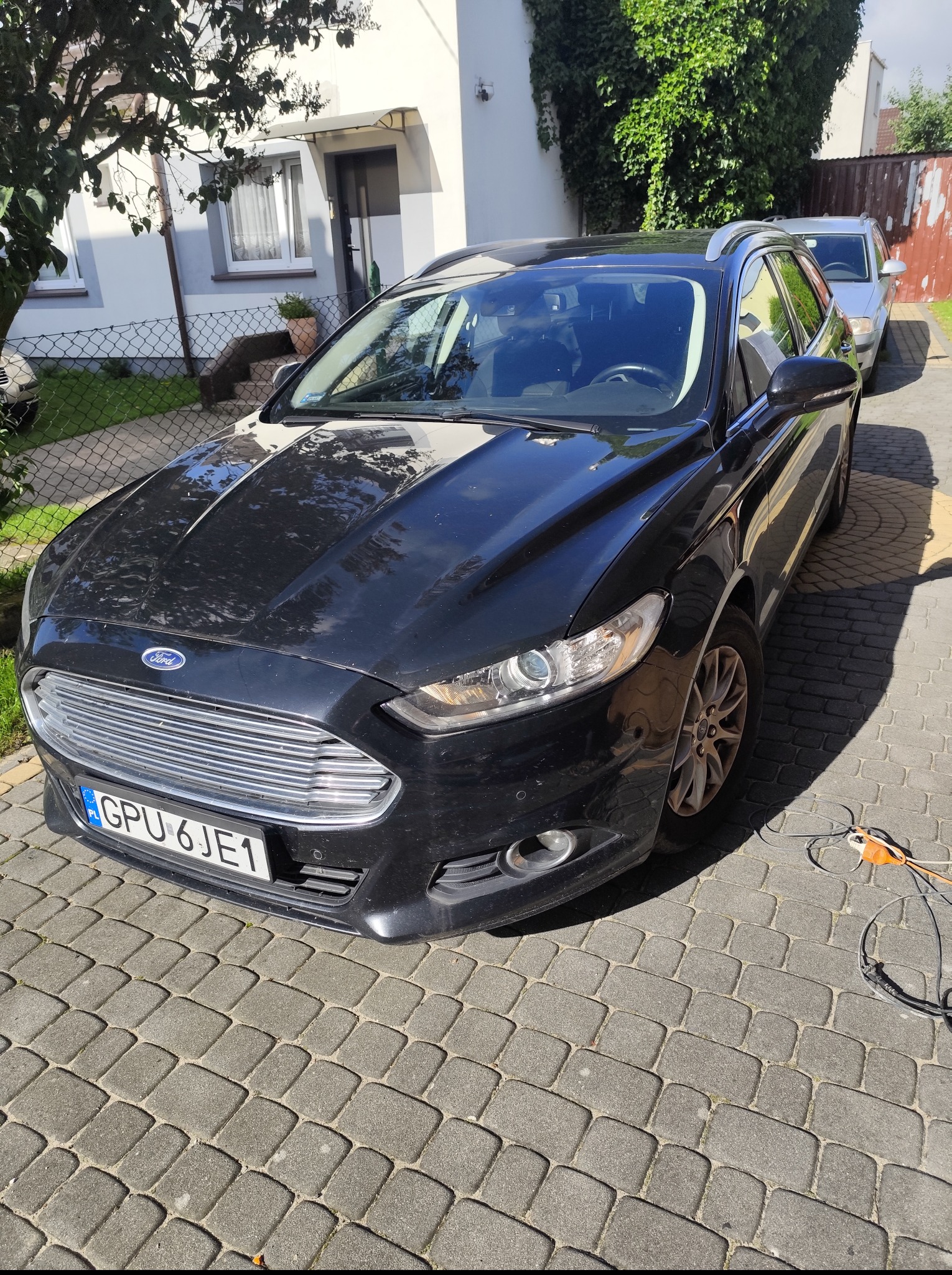 Czarny Ford Mondeo kombi zaparkowany na brukowanym podjeździe przed domem. Widoczny fragment kabla zasilającego, sugerujący prace pielęgnacyjne.