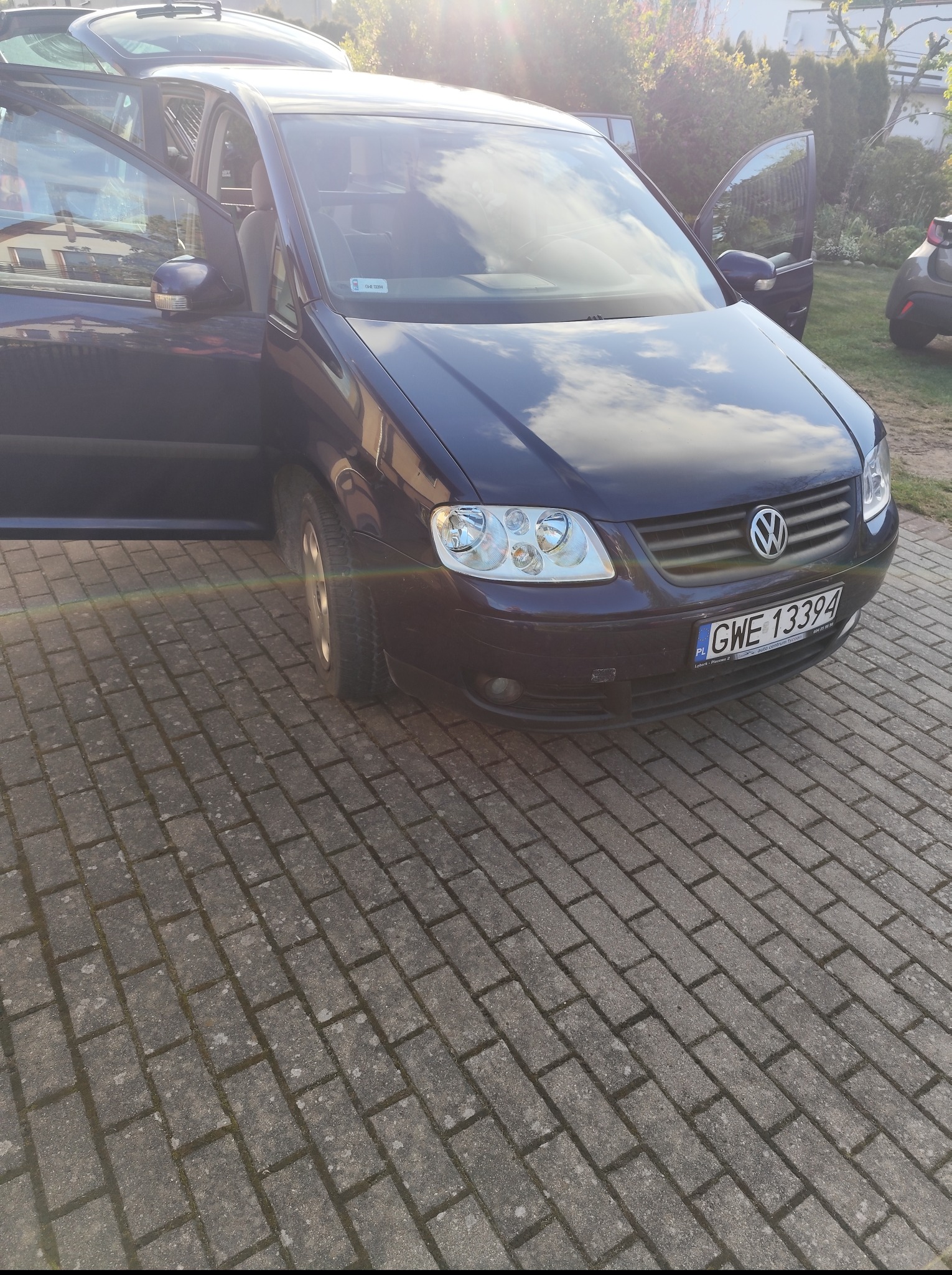 Ciemny minivan Volkswagen Touran z otwartymi drzwiami, zaparkowany na brukowanym podjeździe w słoneczny dzień. Widoczne odbicie chmur na karoserii.