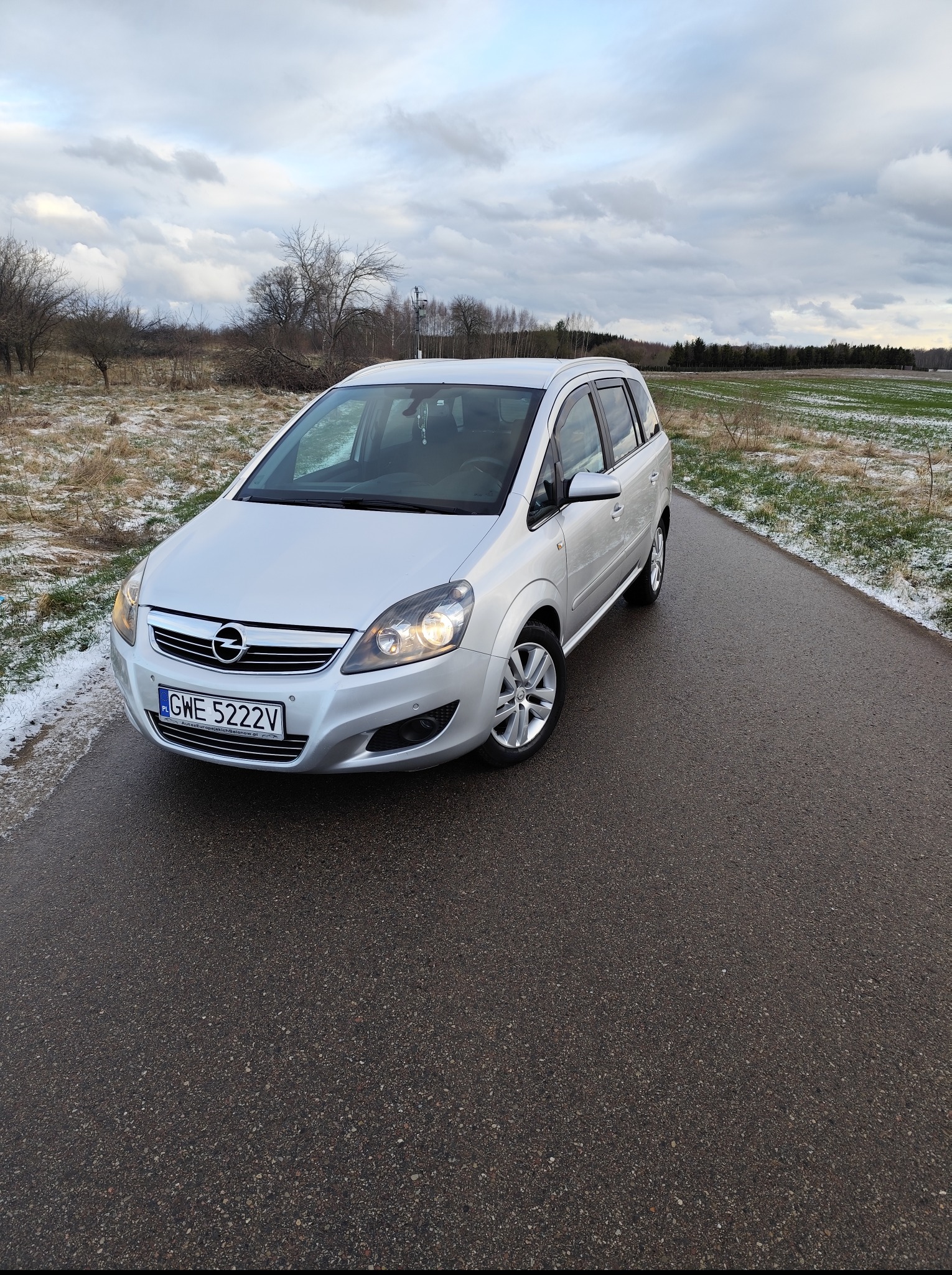 Srebrna Opel Zafira na mokrej, asfaltowej drodze w zimowym krajobrazie. Widoczne włączone światła mijania i tablica rejestracyjna GWE 5222V.