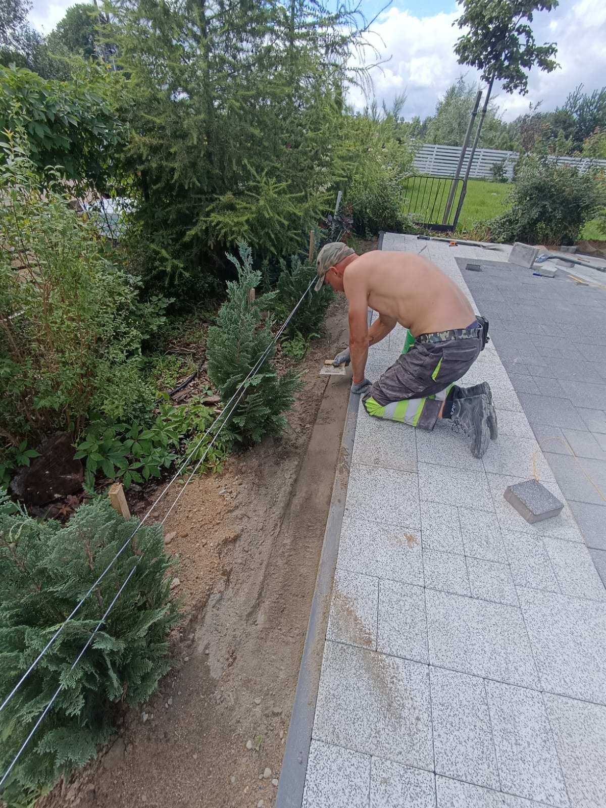 Pracownik w trakcie prac brukarskich: wyrównywanie kostki granitowej przy obrzeżu rabaty w ogrodzie. Widoczne napięte sznurki wyznaczające poziom.