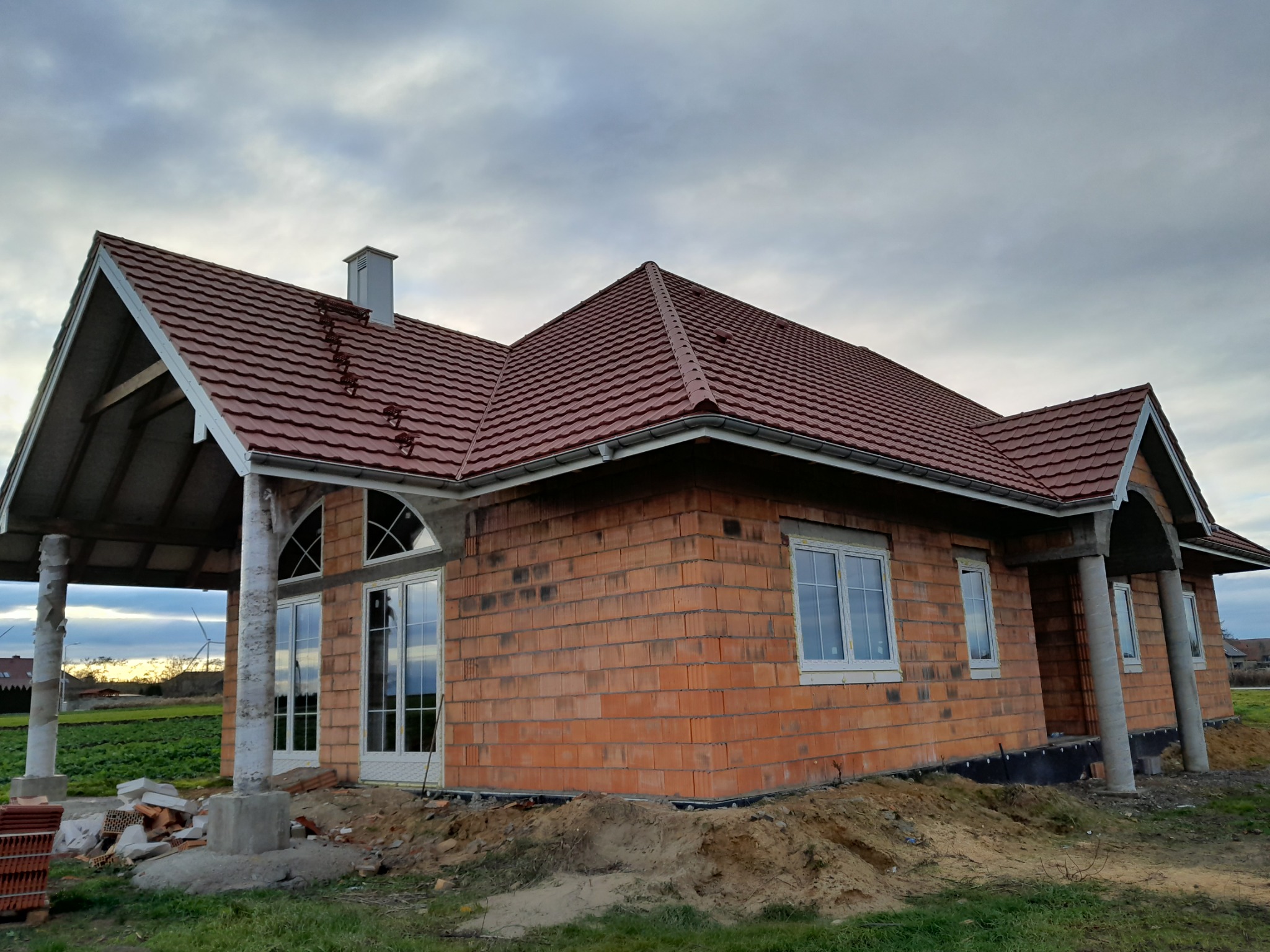 Dom w trakcie budowy z czerwoną dachówką i białymi oknami. Widoczne cegły i kolumny. Pochmurne niebo w tle.