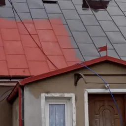 Częściowo pomalowany dach z blachy trapezowej na czerwono, widoczny komin z cegły, fragment rynny i okna. Ujęcie z dołu, widoczne ślady po farbie.