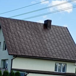 ATADOM SPÓŁKA Z OGRANICZONĄ ODPOWIEDZIALNOŚCIĄ - Dom jednorodzinny z brązowym dachem z blachy trapezowej i ceglanym kominem na tle błękitnego nieba z liniami energetycznymi.