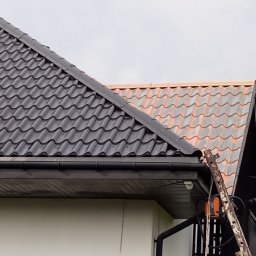 ATADOM SPÓŁKA Z OGRANICZONĄ ODPOWIEDZIALNOŚCIĄ - Fragment dachu dwuspadowego z metalową dachówką w dwóch kolorach (czarny i ceglasty) oraz rynną. Widoczna drabina oparta o rynnę i fragment elewacji budynku.