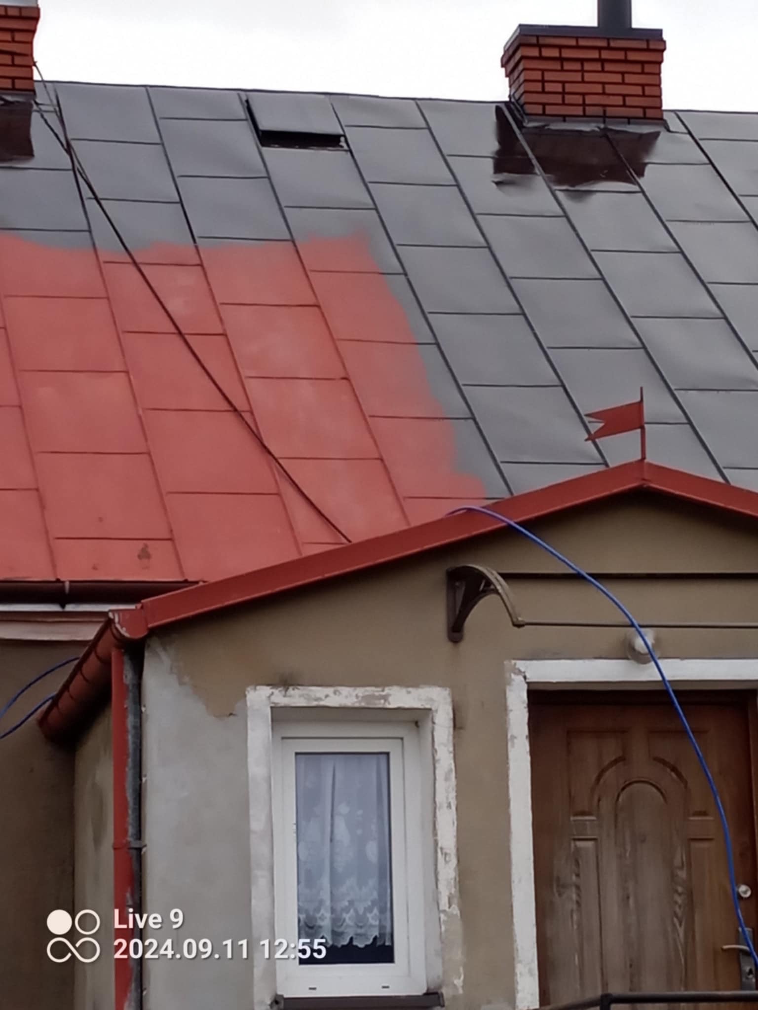 Częściowo pomalowany dach z blachy trapezowej na czerwono, widoczny komin z cegły, fragment rynny i okna. Ujęcie z dołu, widoczne ślady po farbie.