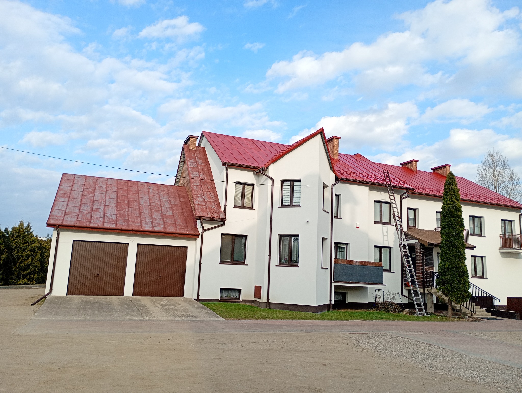 Biały budynek z czerwonym dachem i brązowymi bramami garażowymi. Na dachu drabina. Błękitne niebo z chmurami w tle. Widok z poziomu gruntu.