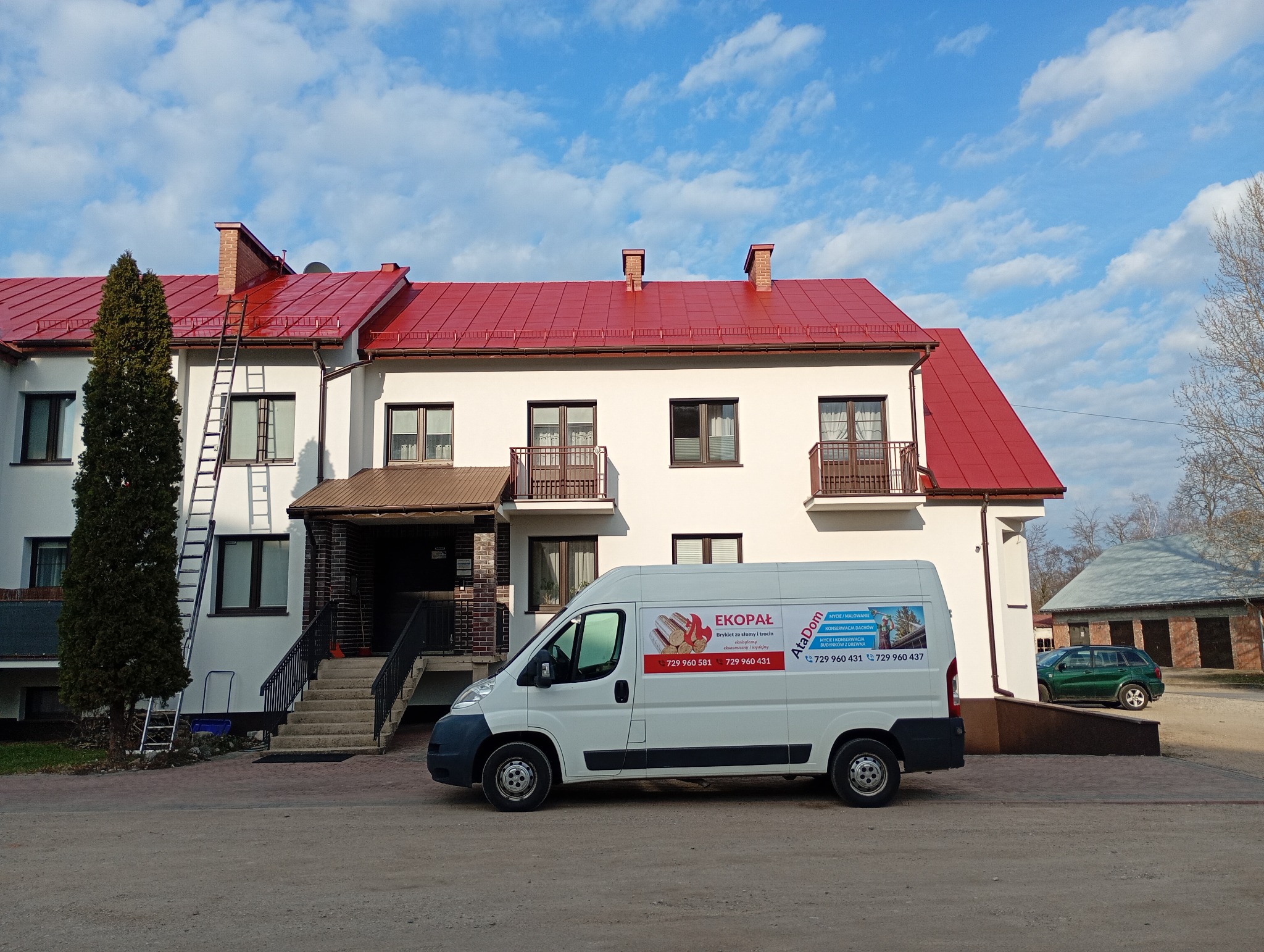 Budynek z czerwonym dachem po renowacji, drabina oparta o ścianę, biały bus z reklamą firmy. Błękitne niebo z chmurami w tle. Widok z poziomu gruntu.