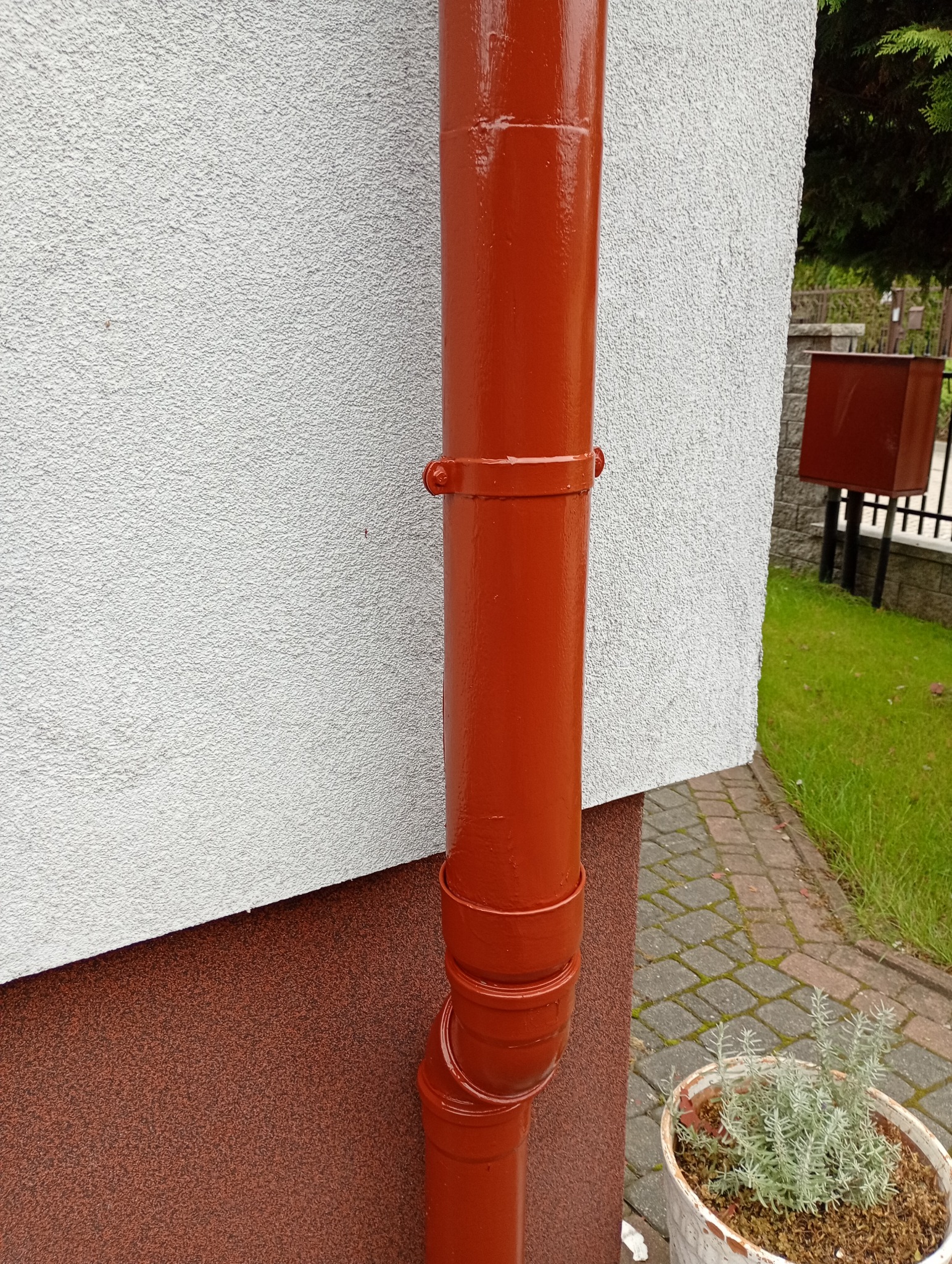 Błyszcząca, pomarańczowa rynna spustowa po renowacji, przymocowana do białej ściany budynku. W tle widoczny fragment trawnika i kostka brukowa.