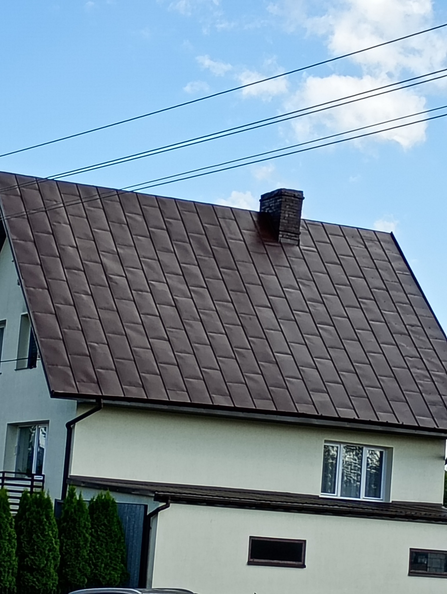 Dom jednorodzinny z brązowym dachem z blachy trapezowej i ceglanym kominem na tle błękitnego nieba z liniami energetycznymi.