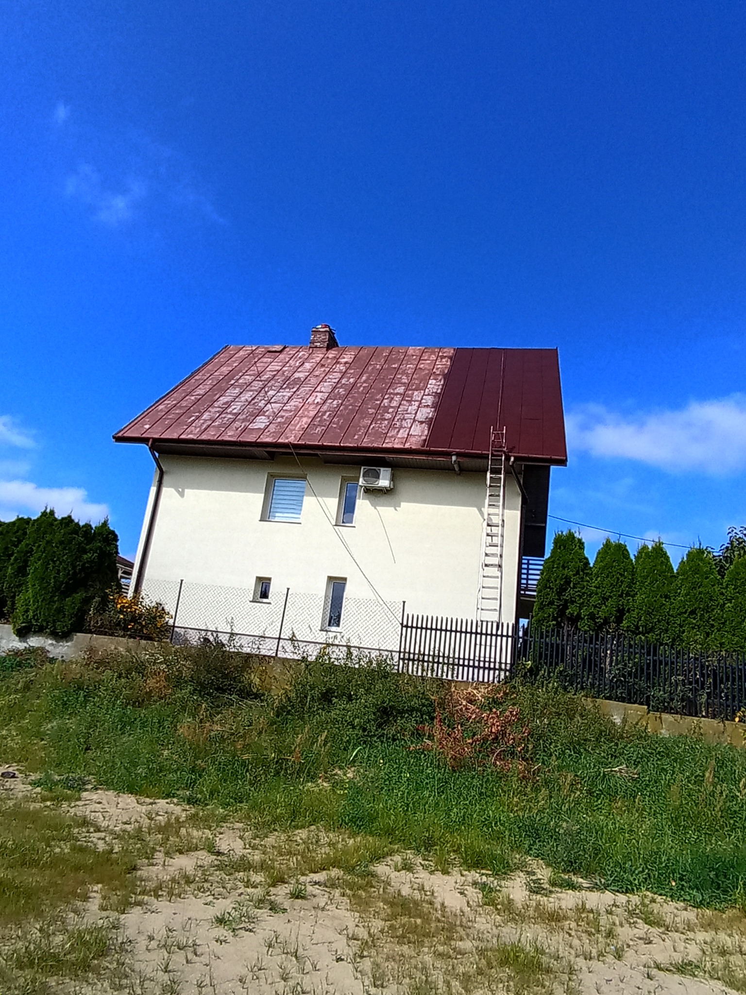 Dom z częściowo pomalowanym na ciemny brąz dachem, drabina oparta o ścianę. Widoczny kontrast między starą a nową powłoką. Błękitne niebo w tle.
