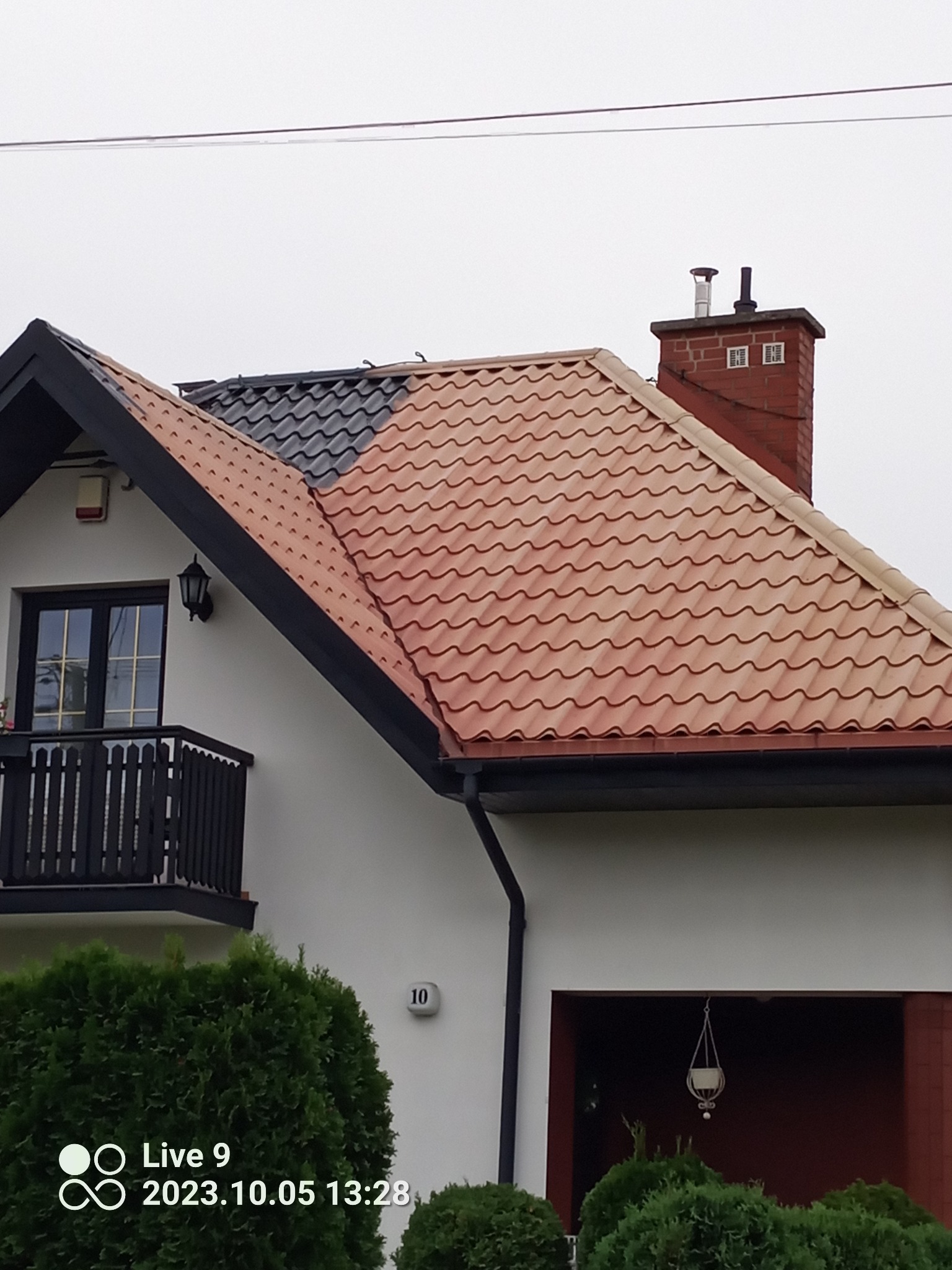 Częściowo odnowiony dach z dachówką ceramiczną i komin na tle nieba w Andrzejki-Tyszki. Widoczna część dachu w kolorze czarnym i część w kolorze czerwonym.