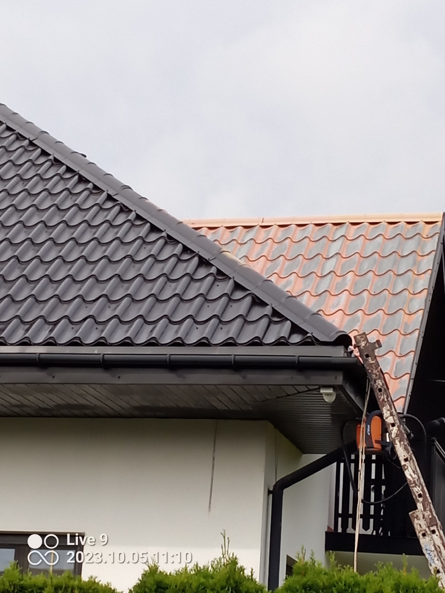 Fragment dachu dwuspadowego z metalową dachówką w dwóch kolorach (czarny i ceglasty) oraz rynną. Widoczna drabina oparta o rynnę i fragment elewacji budynku.