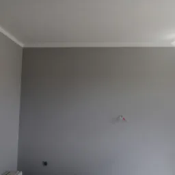 Ściana pomalowana na szaro z białą listwą sufitową w Olkuszu. Widoczne gniazdko elektryczne i wystające przewody. Świeżo po malowaniu.