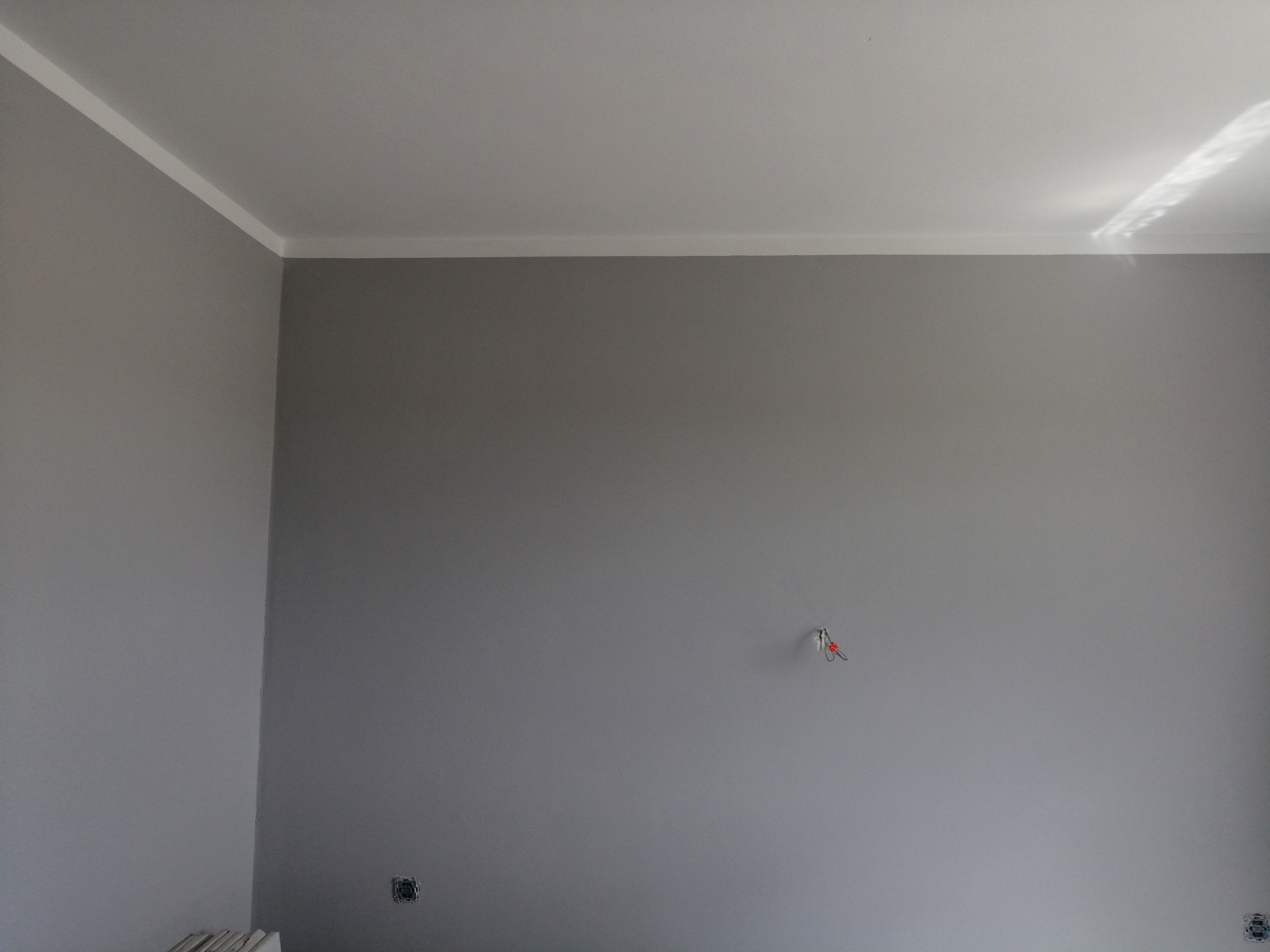 Ściana pomalowana na szaro z białą listwą sufitową w Olkuszu. Widoczne gniazdko elektryczne i wystające przewody. Świeżo po malowaniu.