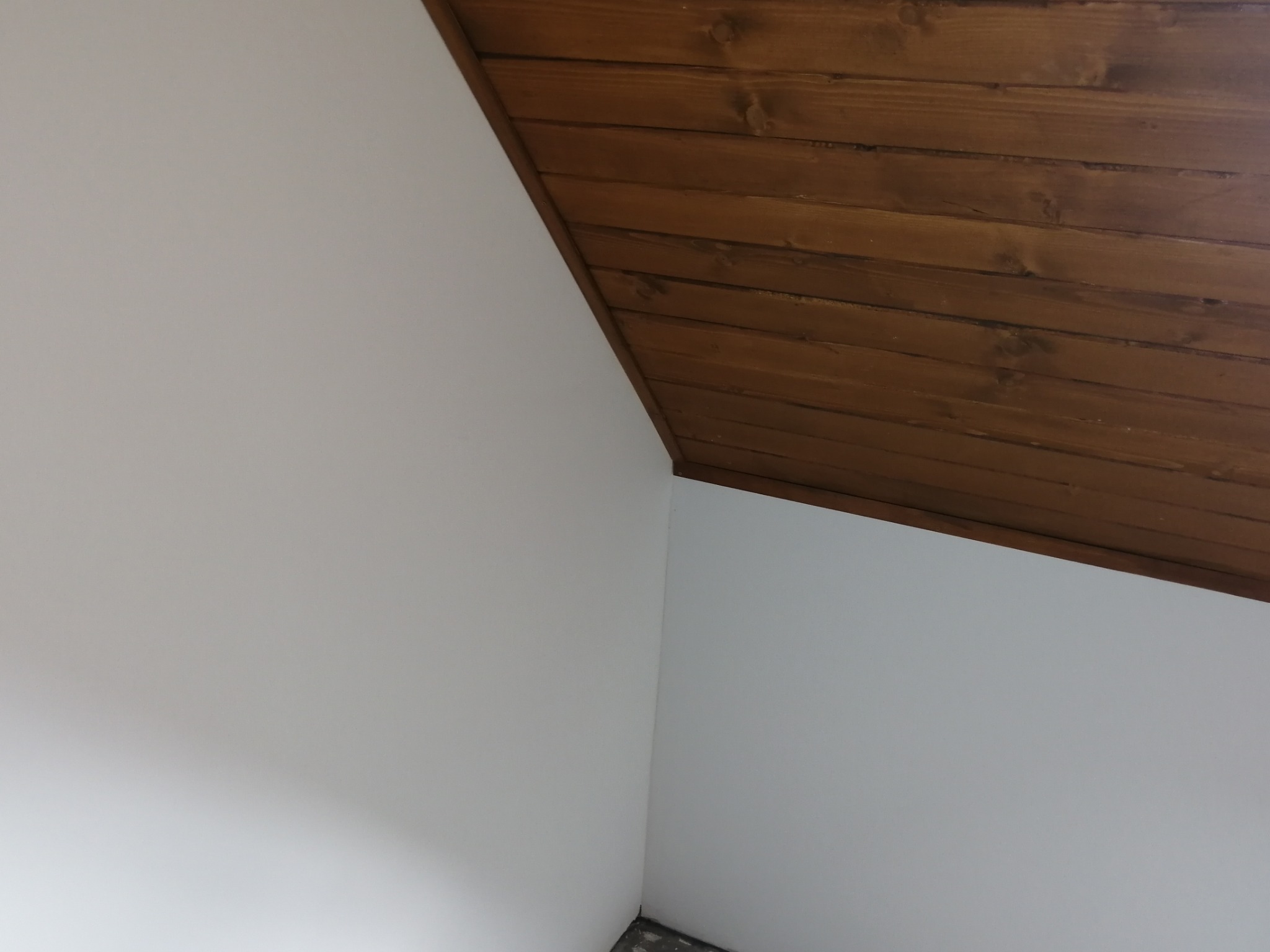 Ściana pomalowana na biało łącznie z sufitem w rogu pokoju. Część sufitu wykończona brązowymi panelami drewnianymi.