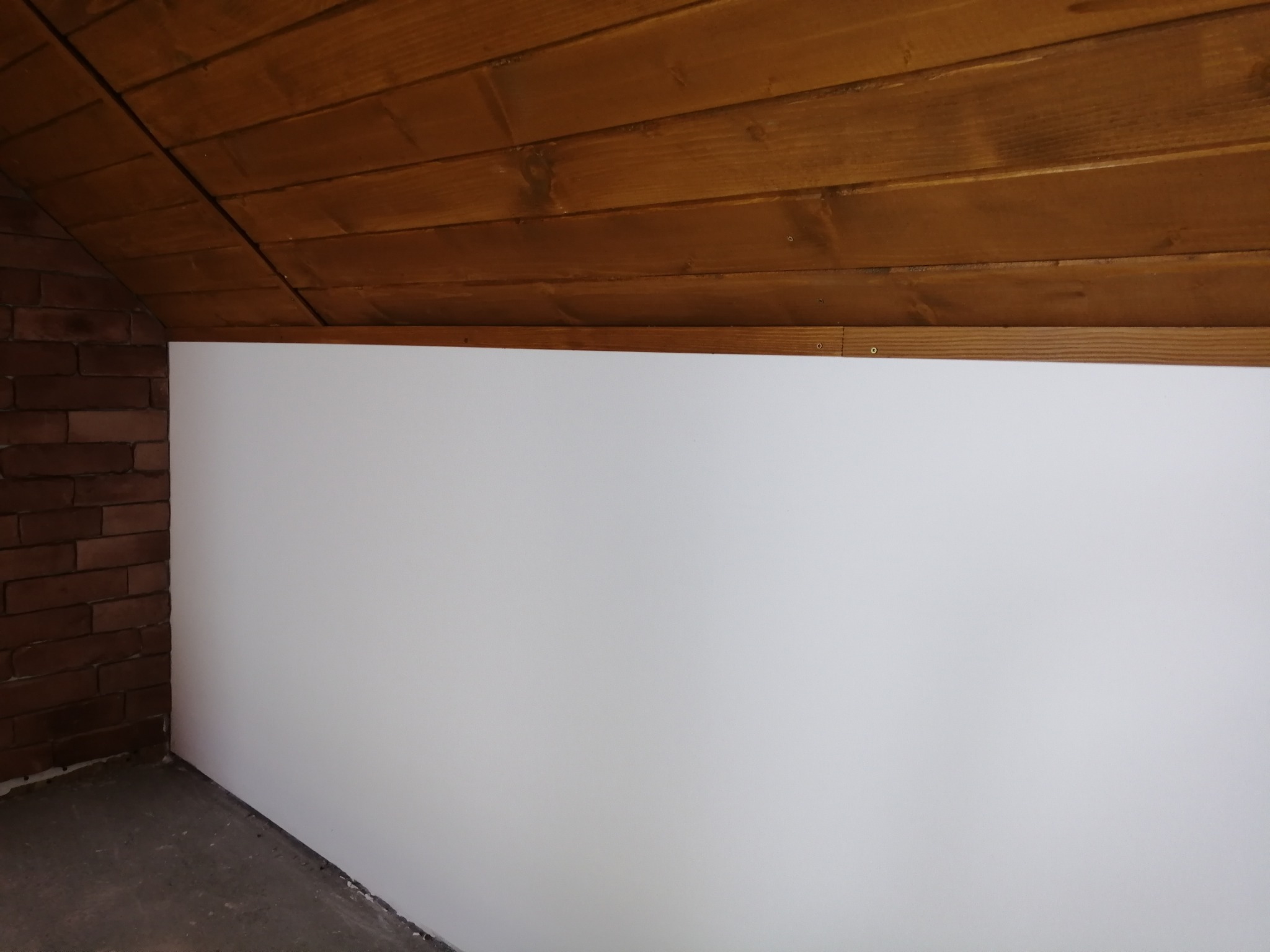 Biała ściana w pomieszczeniu z drewnianym sufitem i fragmentem ceglanej ściany. Widoczne łączenie sufitu ze ścianą. Kąt ujęcia z dołu.