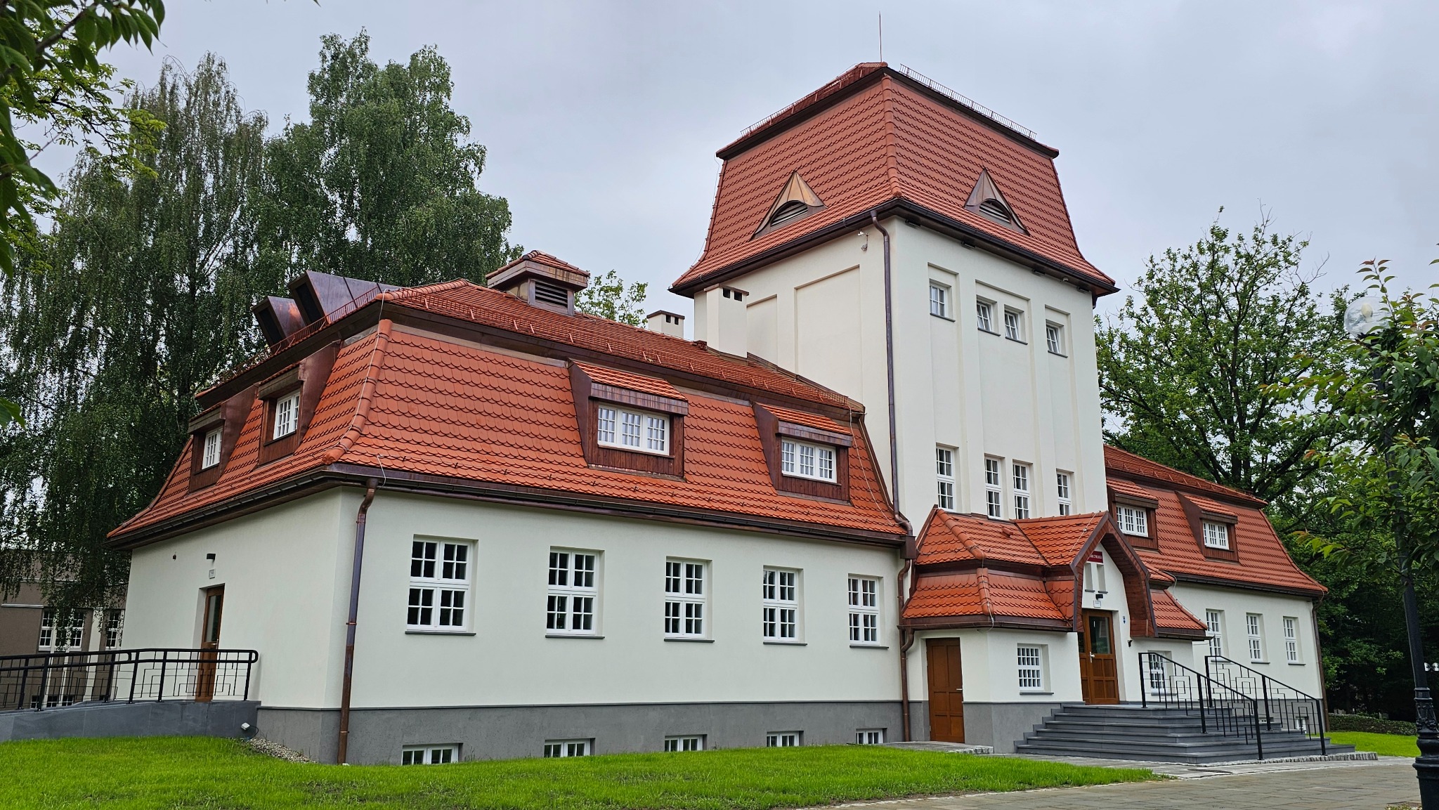 Elewacja budynku z czerwoną dachówką, widok z zewnątrz. Jasna fasada, regularne okna, dwuspadowy dach i wieża. Otoczenie zielone, drzewa w tle.