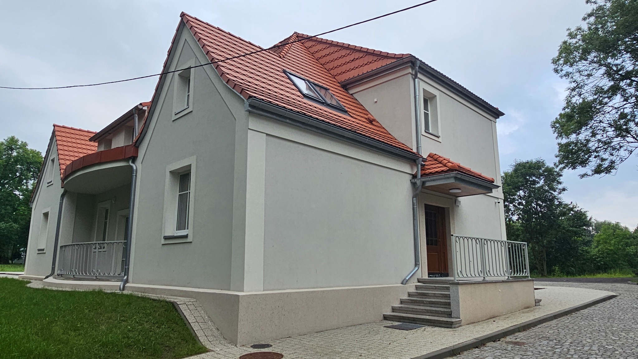 Elewacja domu z czerwoną dachówką, widok z boku. Jasna fasada, białe okna, schody wejściowe. Widoczne drzewa w tle. Ujęcie bez filtrów, naturalne oświetlenie.