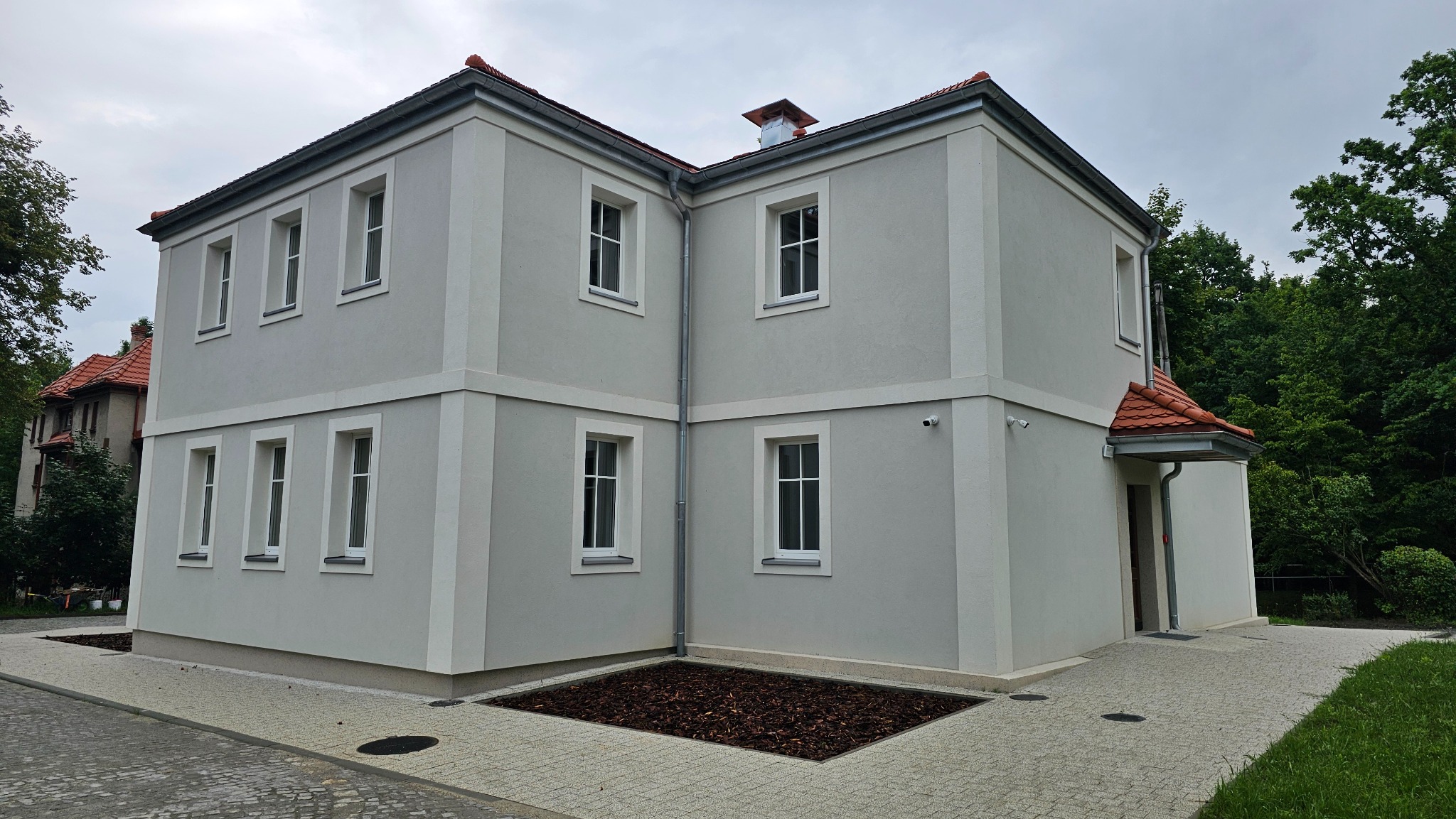 Odnowiona elewacja dwupiętrowego budynku z szarym tynkiem, białymi obramowaniami okien i czerwoną dachówką. Widoczny fragment chodnika z kostki brukowej i rabata.