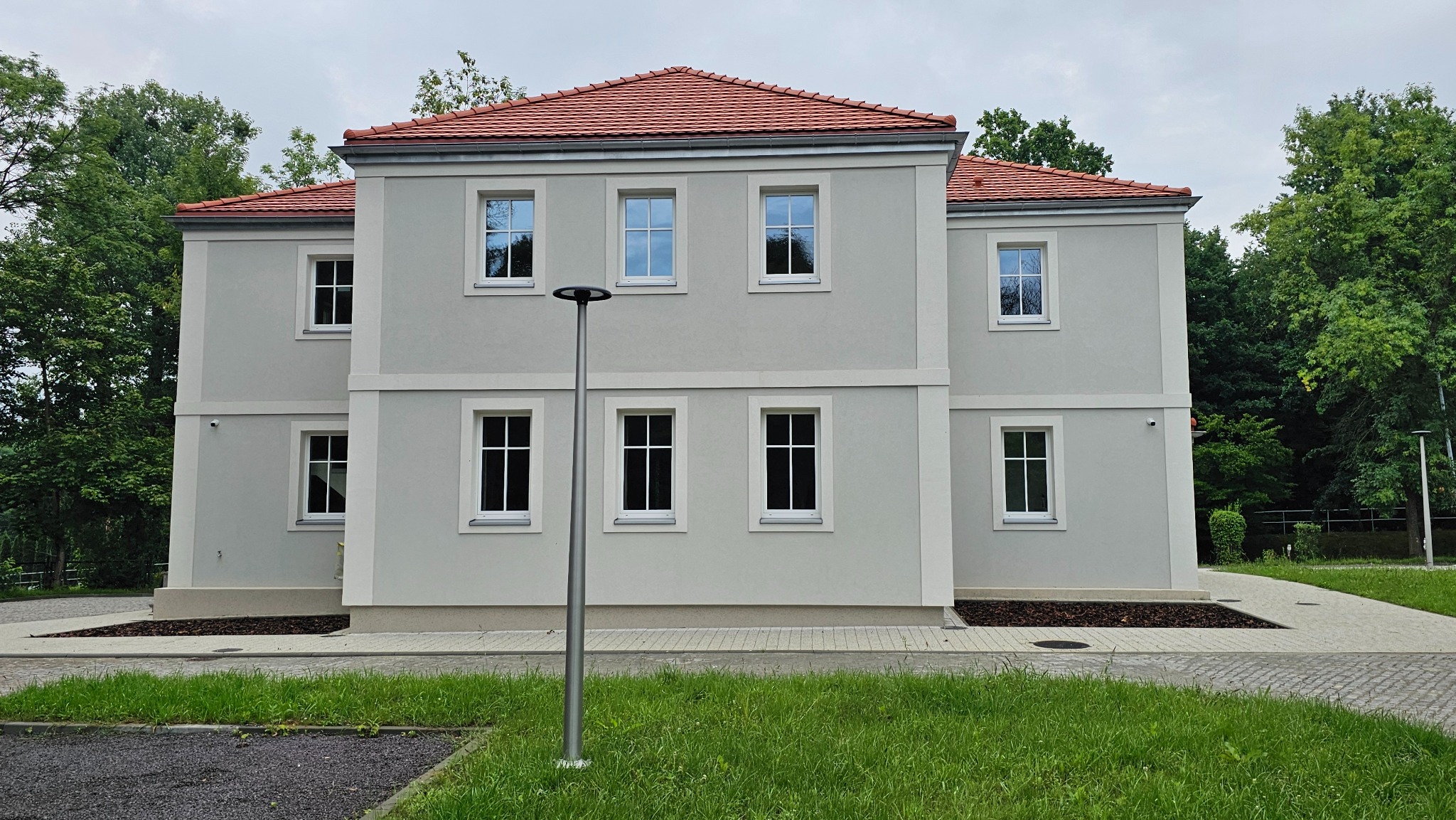 Dwukondygnacyjny dom z szarą elewacją, czerwoną dachówką i białymi obramowaniami okien, widziany z poziomu trawnika w słoneczny dzień.