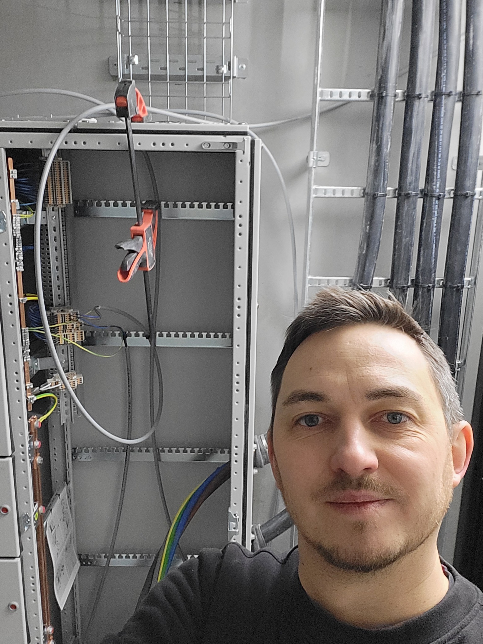 Elektryk prezentuje rozdzielnicę z okablowaniem w trakcie montażu. Widoczne przewody i kable w szarym panelu oraz narzędzia monterskie.