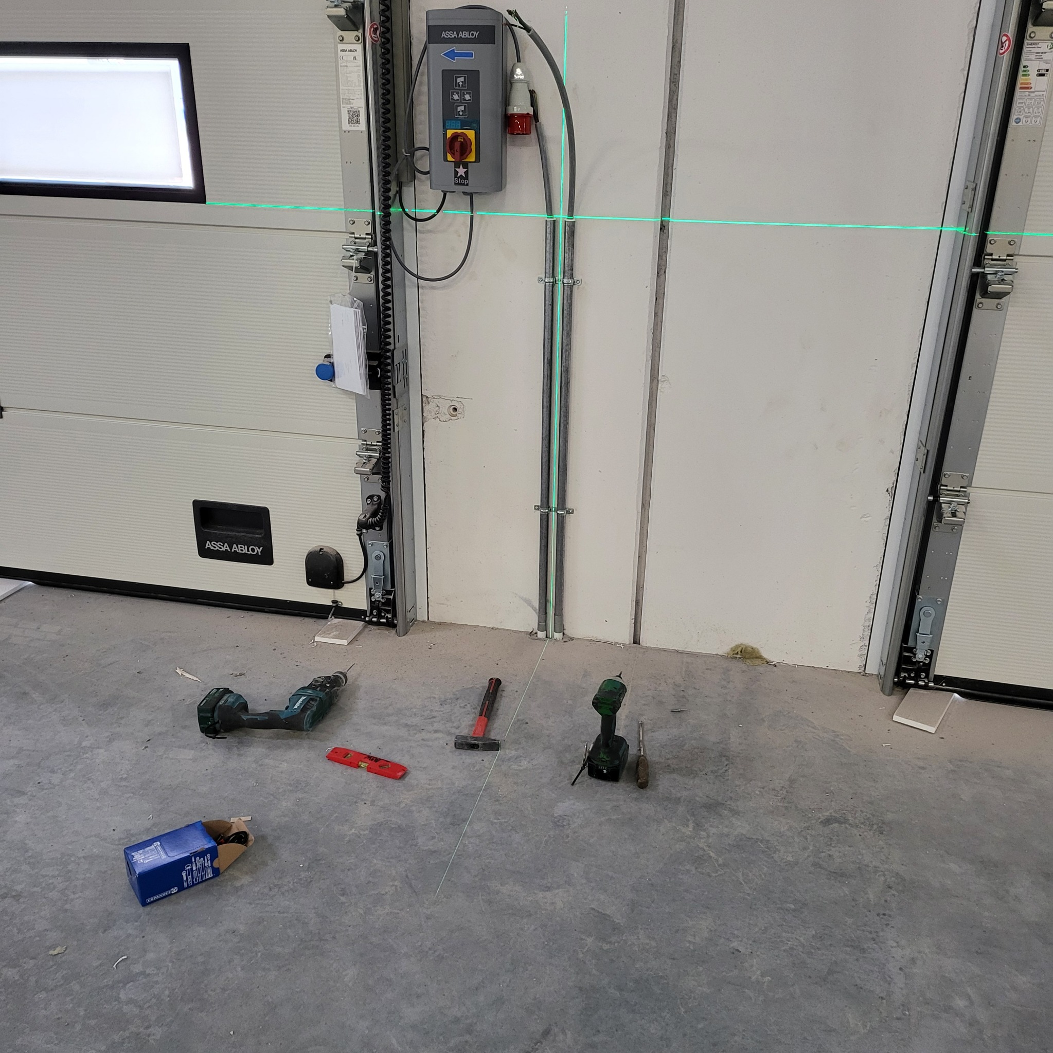 Montaż elektryki do bramy garażowej: widoczny panel sterowania, przewody w peszlu i narzędzia pracy, w tym poziomica laserowa.