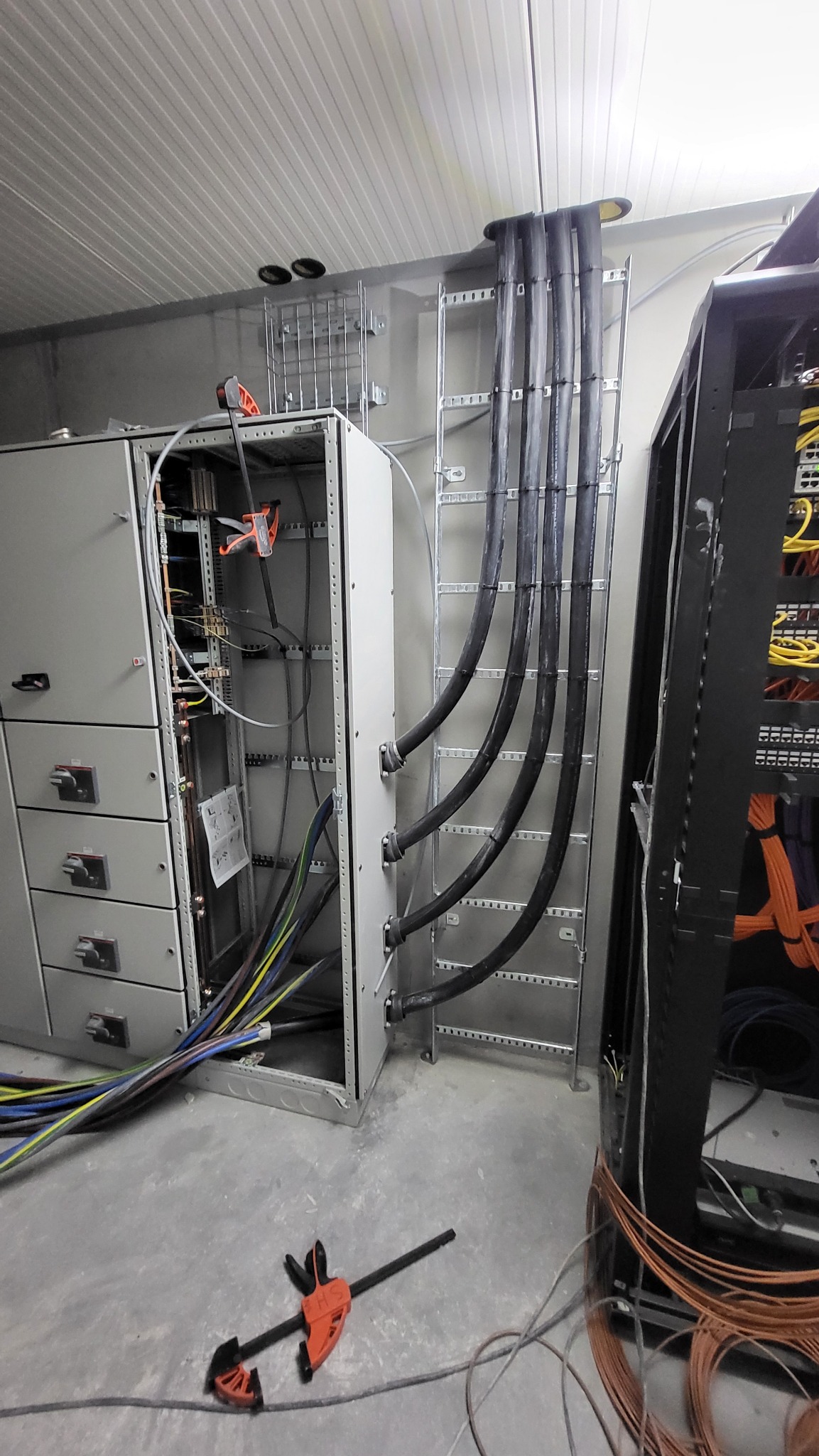 Rozdzielnia elektryczna w szarej obudowie z otwartymi drzwiami, widoczne kable w peszlach, część z nich zamocowana na drabince kablowej.