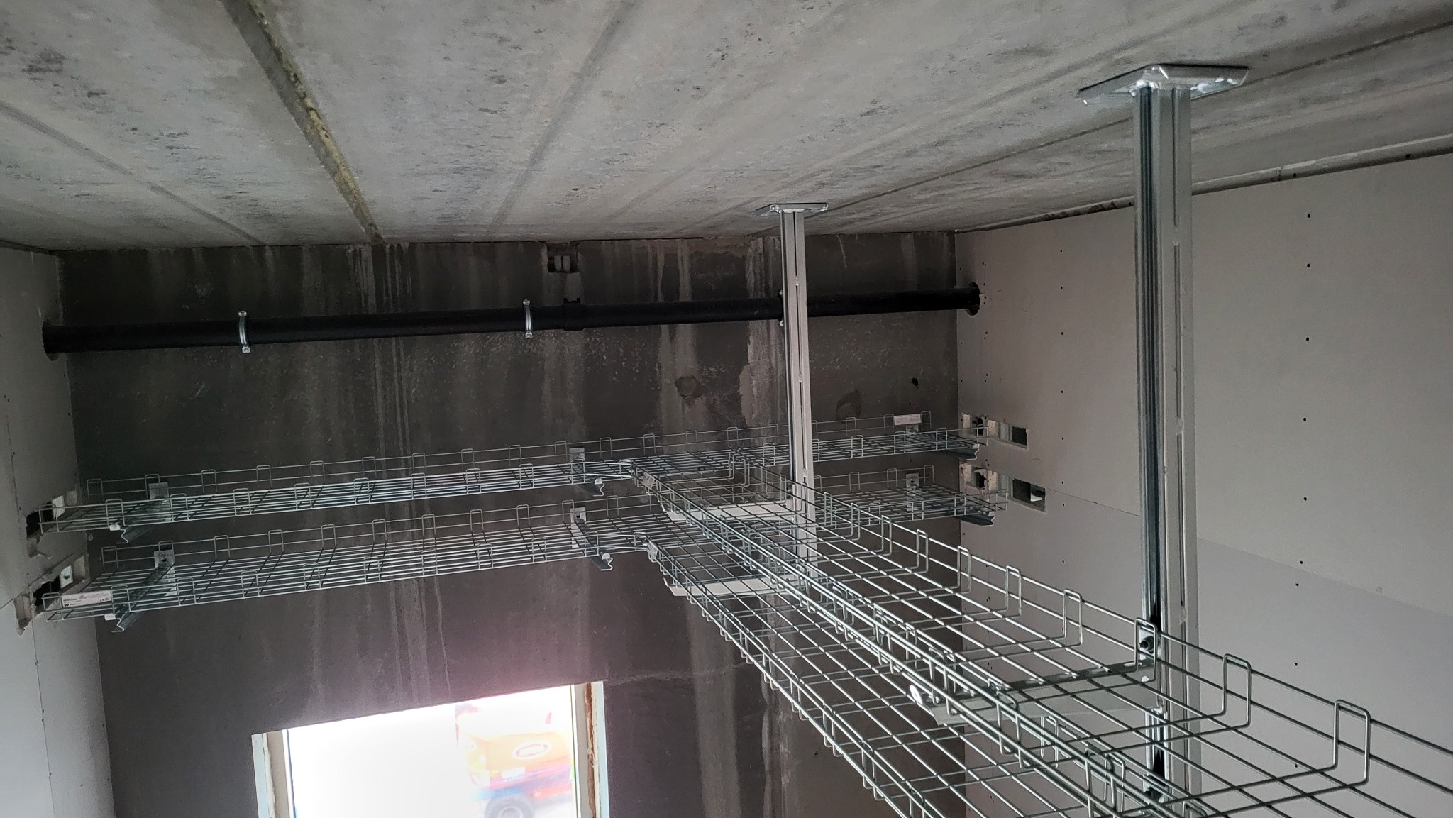 Metalowe korytka kablowe zamontowane pod sufitem w surowym pomieszczeniu, widoczne rury i elementy konstrukcyjne. Kąt ujęcia z dołu podkreśla perspektywę instalacji.