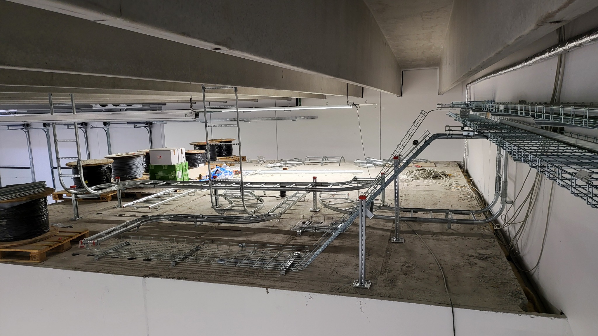 Montaż tras kablowych pod sufitem w hali przemysłowej. Widoczne kable na szpulach, drabinki kablowe i elementy mocujące. Surowy stan wykończenia.