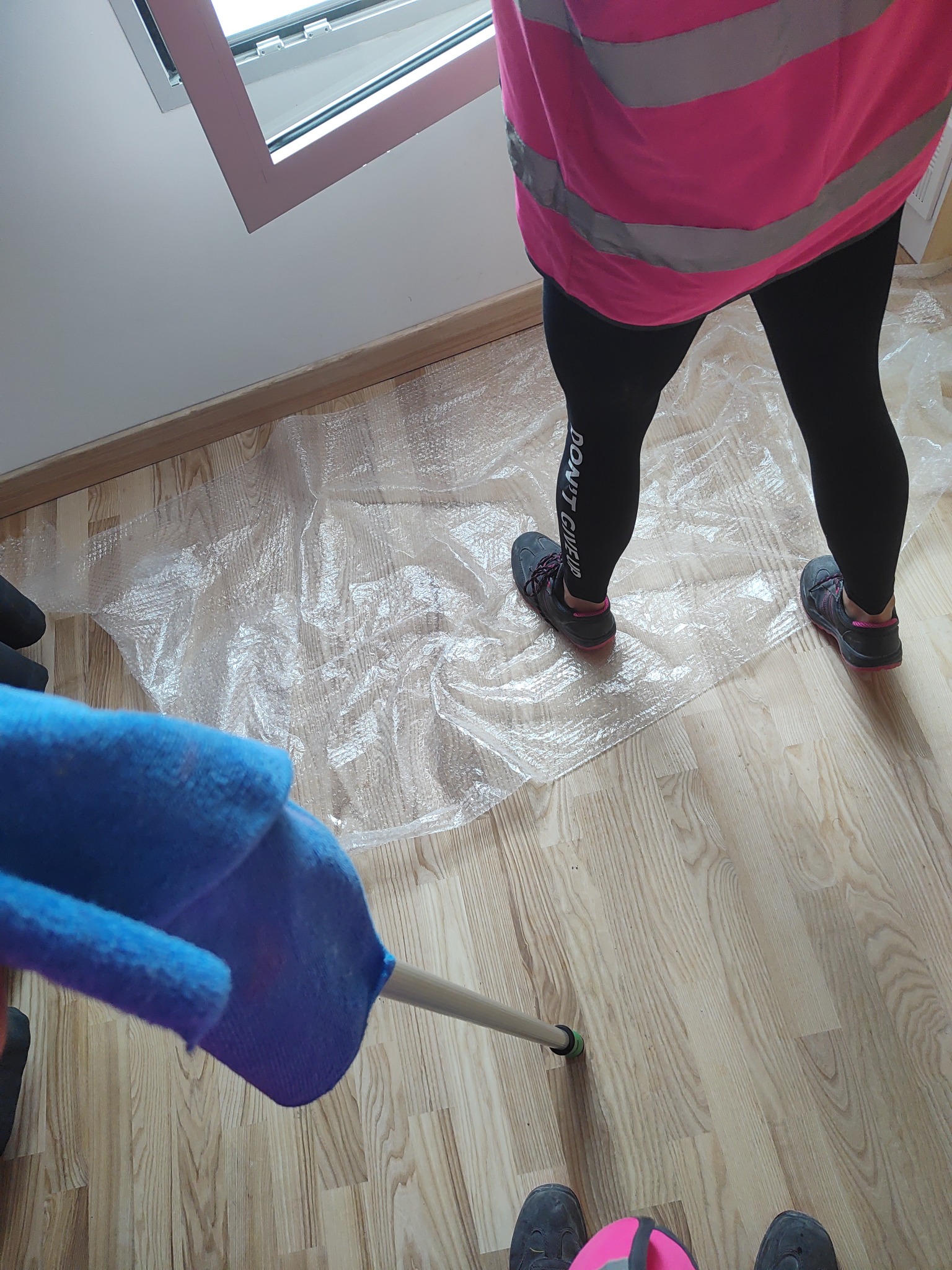 Osoba w odblaskowej kamizelce czyści podłogę mopem, zabezpieczoną folią bąbelkową, widoczne fragmenty okna i podłogi.