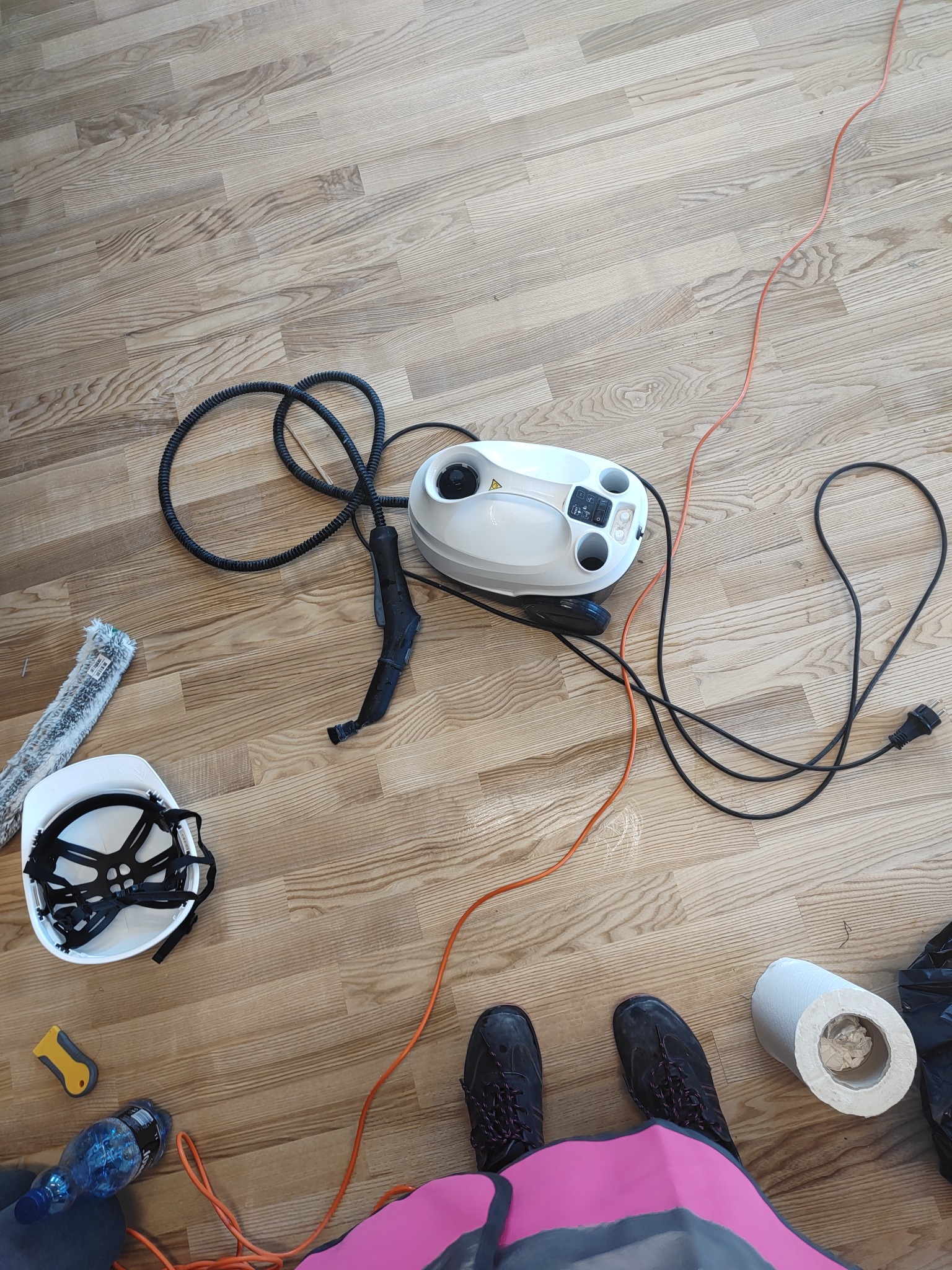 Parownica, ściągaczka do okien, kask i inne akcesoria do sprzątania na podłodze z paneli. Widoczne buty osoby wykonującej usługę.