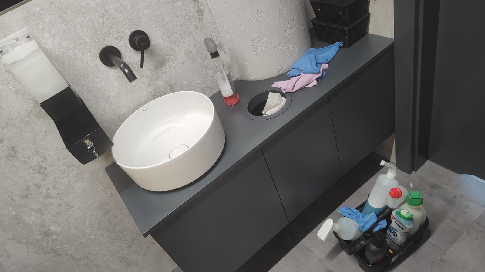 Umywalka z czarną armaturą i dozownikiem mydła. Obok środki czystości i ściereczki, sugerujące świeżo posprzątane pomieszczenie z elementami wyposażenia.
