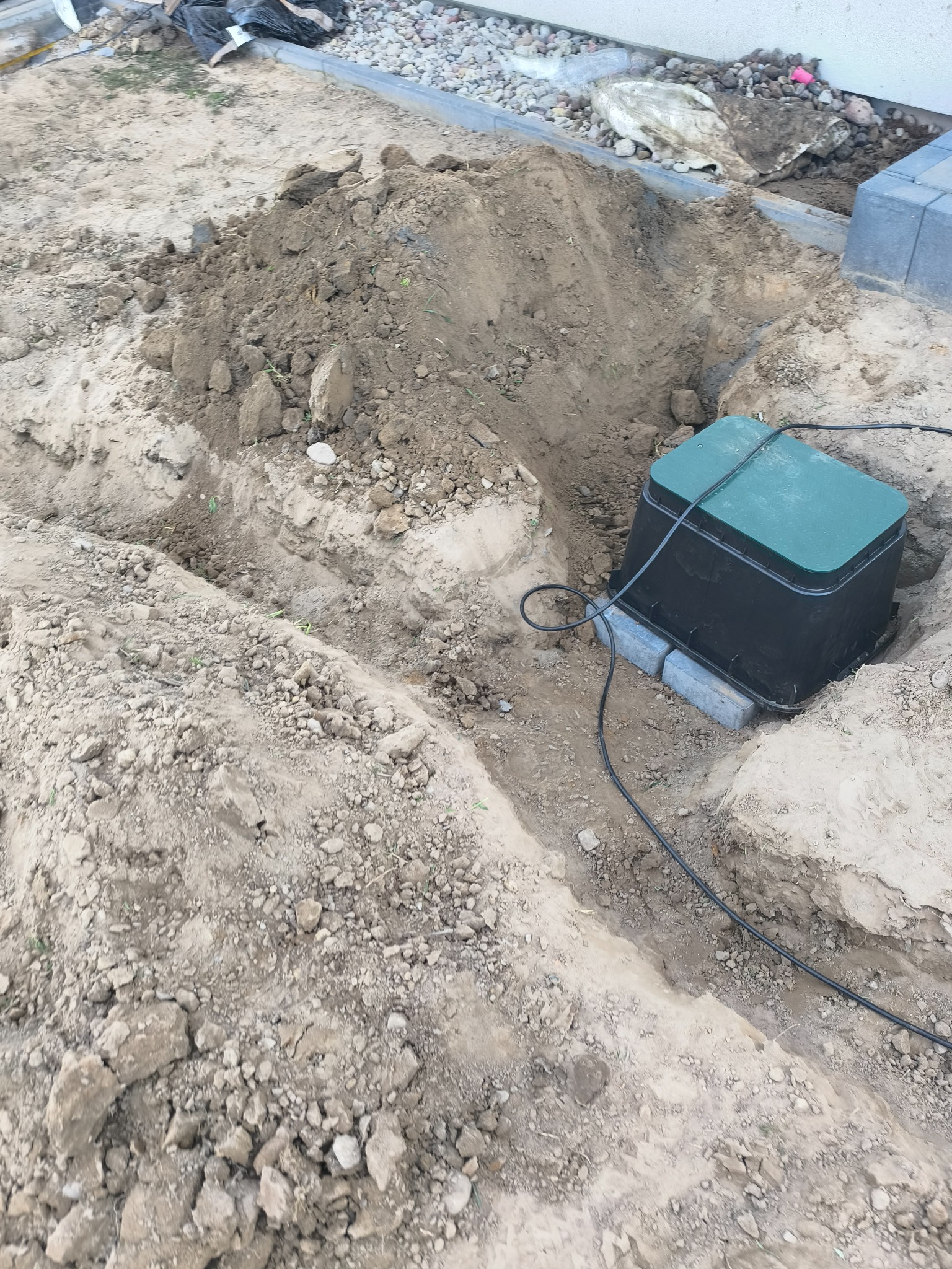 Instalacja podziemnego systemu nawadniania: wykopane rowy, skrzynka sterująca z zieloną pokrywą i kable. Etap prac w ogrodzie.