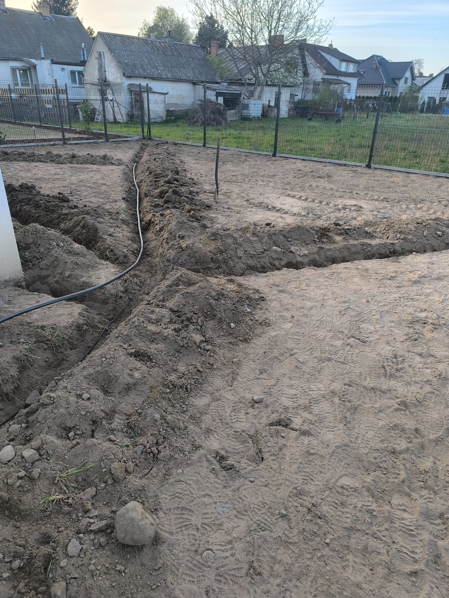 Przygotowanie terenu pod ogród: wykopane rowy, widoczny przewód instalacji nawadniającej. Tło: domy jednorodzinne i ogrodzenie z siatki.