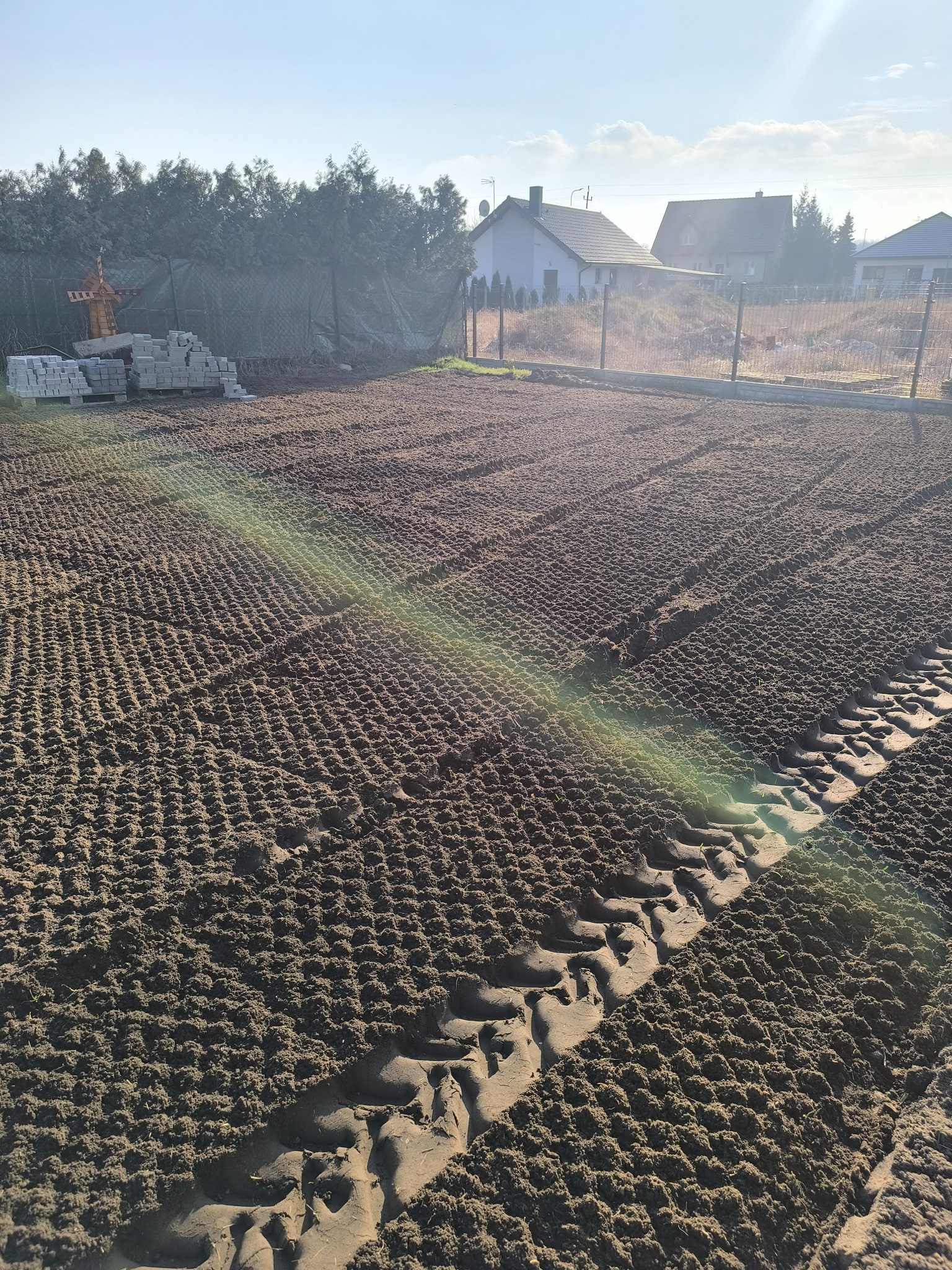 Przygotowane podłoże pod trawnik z widocznymi śladami maszyn i ułożonymi obrzeżami. W tle domy i drzewa.