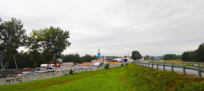 Szeroki kadr na skład budowlany z betonowymi elementami, ciężarówkami i wieżą do mieszania. Widok zza barierek, z zielonym trawnikiem i drzewami po bokach.