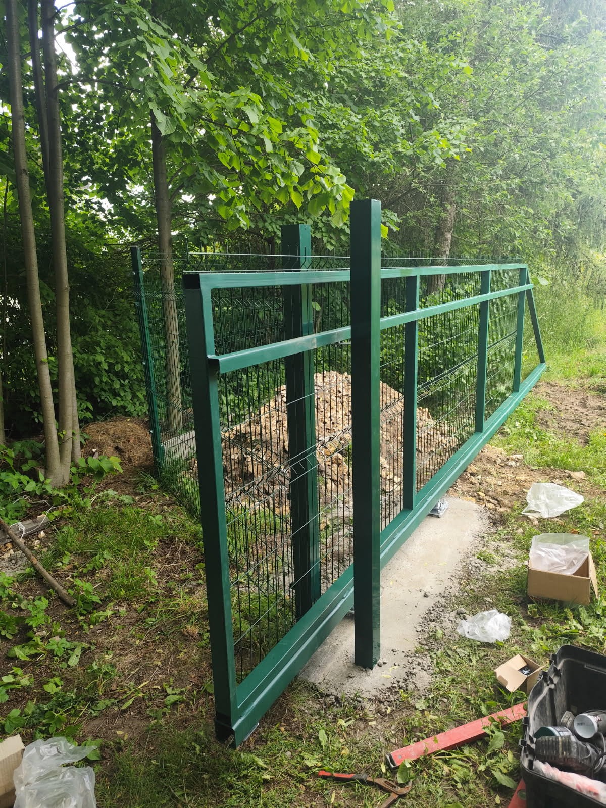Zielona, metalowa brama panelowa w trakcie montażu, widoczne słupki w betonie i narzędzia. Tło: zieleń drzew i krzewów, częściowo zamaskowana siatką.