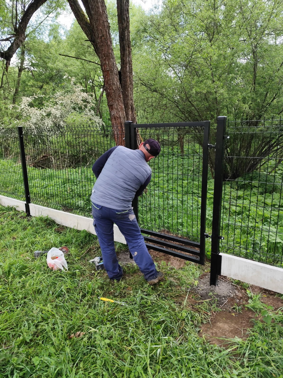 Instalacja czarnej bramki w ogrodzeniu panelowym na trawiastym terenie. Widoczny monter w roboczym ubraniu przy pracy, z narzędziami obok. Tło: drzewa i krzewy.