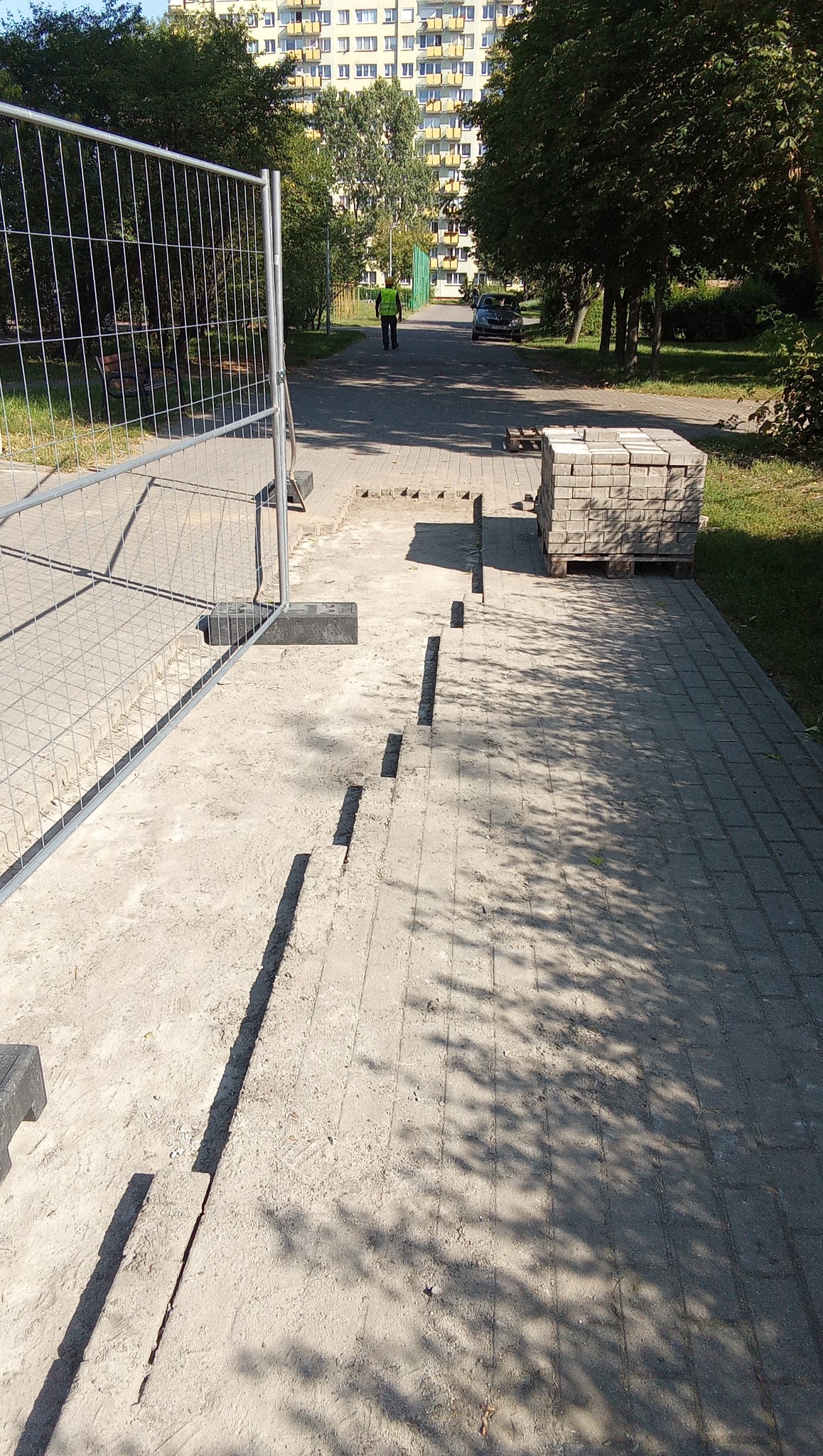 Fragment chodnika z wyciętymi szczelinami pod kostkę brukową, zabezpieczony metalowym ogrodzeniem. W tle blok i osoba w kamizelce odblaskowej.