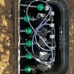 Łukasz Witak - Czarna skrzynka z zaworami i przewodami systemu nawadniania, widok z góry. Elementy złączek w kolorze zielonym. Skrzynka umieszczona w ziemi.