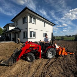 Łukasz Witak - Czerwony traktor z glebogryzarką przygotowuje teren pod ogród na tle nowoczesnego domu z czarnym SUVem i błękitnego nieba z białymi chmurami.