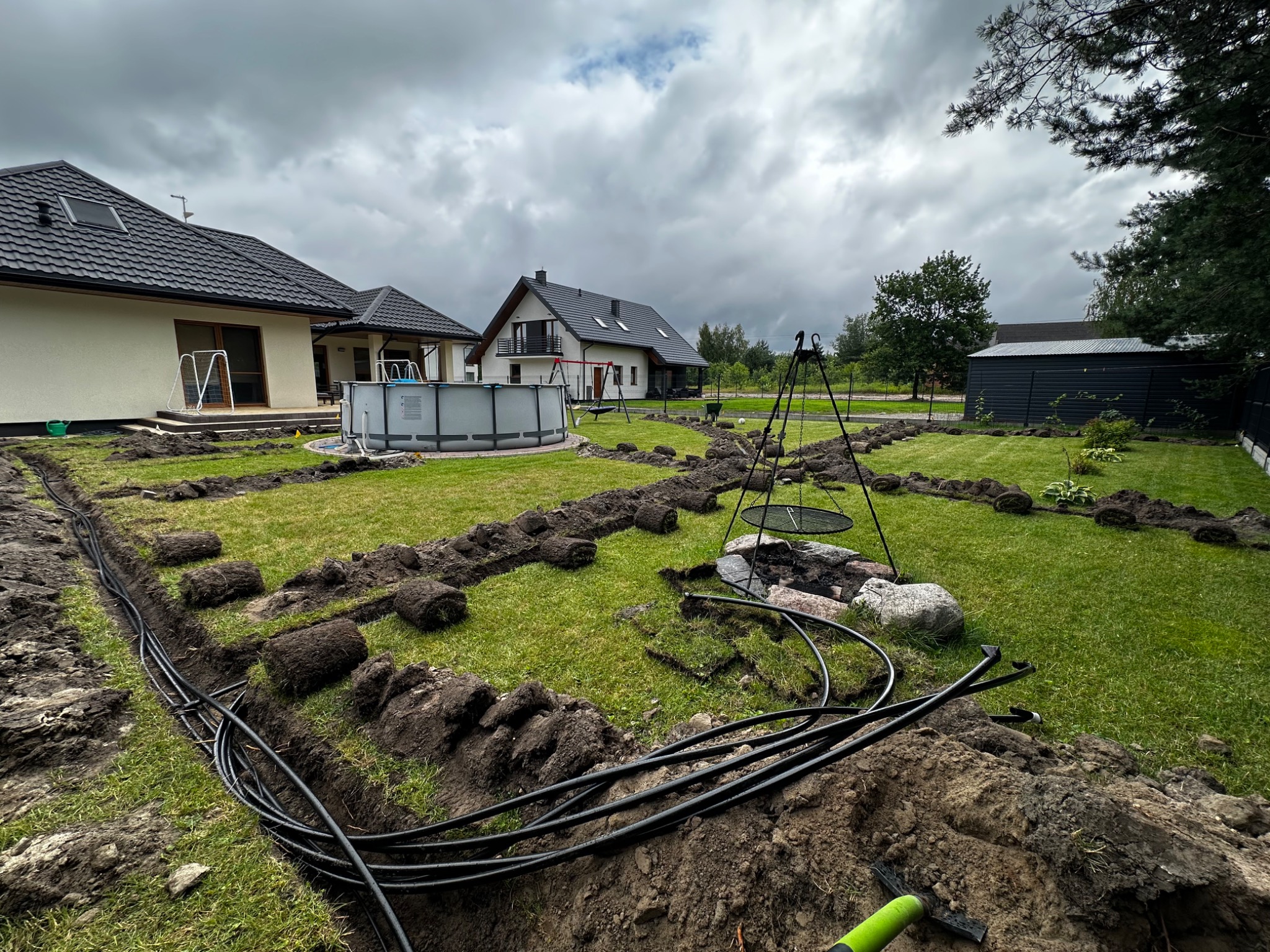 Instalacja nawadniania w ogrodzie: wykopane rowy, czarne rury, odłożone pasy darni. W tle domy, basen i grill na trójnogu pod pochmurnym niebem.