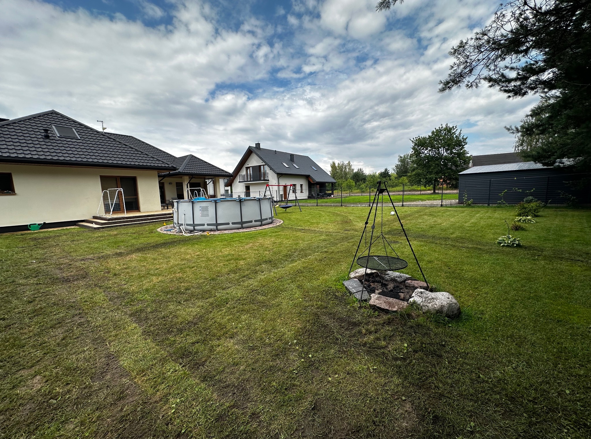 Rozległy ogród z basenem i miejscem na ognisko w Osiecku. Widoczny trawnik po aeracji, domy w tle, pochmurne niebo. Elementy dekoracyjne i rekreacyjne.