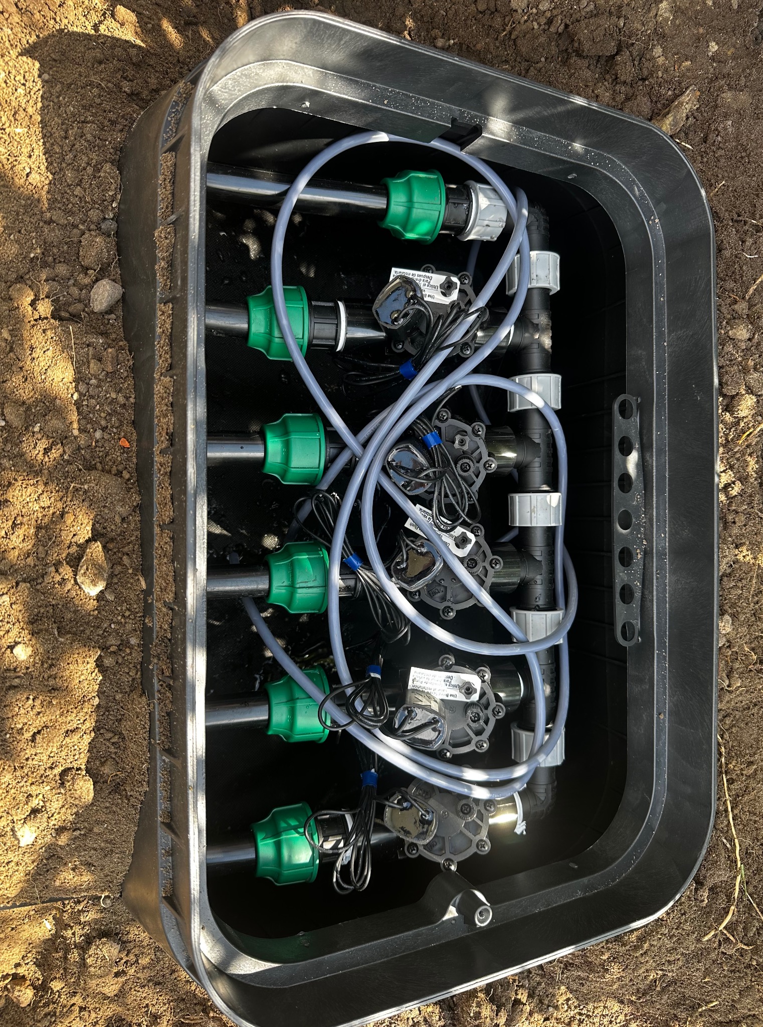 Czarna skrzynka z zaworami i przewodami systemu nawadniania, widok z góry. Elementy złączek w kolorze zielonym. Skrzynka umieszczona w ziemi.