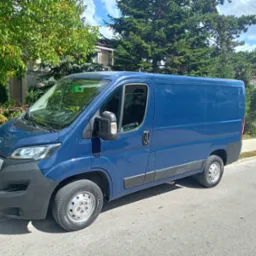 Niebieski bus dostawczy Peugeot zaparkowany na ulicy, z zielenią drzew w tle i błękitnym niebem z chmurami. Widok z boku, w słoneczny dzień.