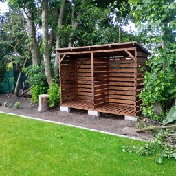 Fjordis - Drewniana, brązowa wiata na drewno z ażurowymi ścianami, umieszczona w ogrodzie na betonowych bloczkach. W tle drzewa i trawa.