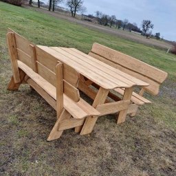 Fjordis - Drewniany stół piknikowy z ławkami, z surowego drewna, ustawiony na trawie, z drzewami w tle. Widok z boku, pochmurne niebo.