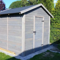 Szara, drewniana budka narzędziowa z czarnym dachem, umieszczona na kostce brukowej w ogrodzie. Widoczne krzewy i drzewa w tle.