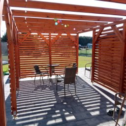 Fjordis - Drewniana pergola z dekoracyjnymi lampionami i meblami ogrodowymi na brukowanym tarasie, tworząca zacienioną strefę relaksu.